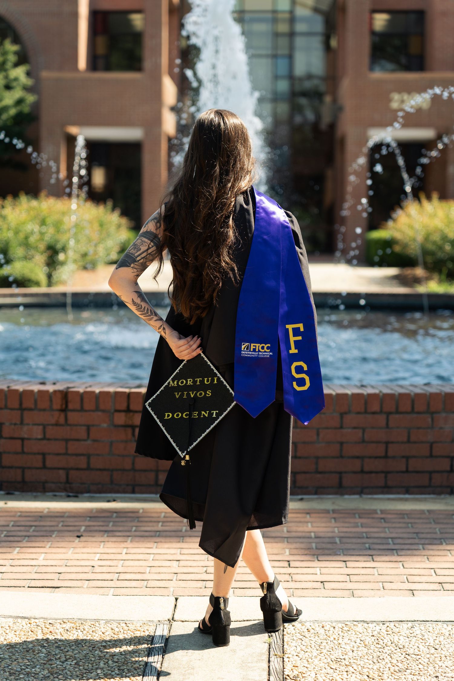 Graduate in black gown with blue FRCC stole holding cap reading 'Mortui Vivos Docent' by a fountain.