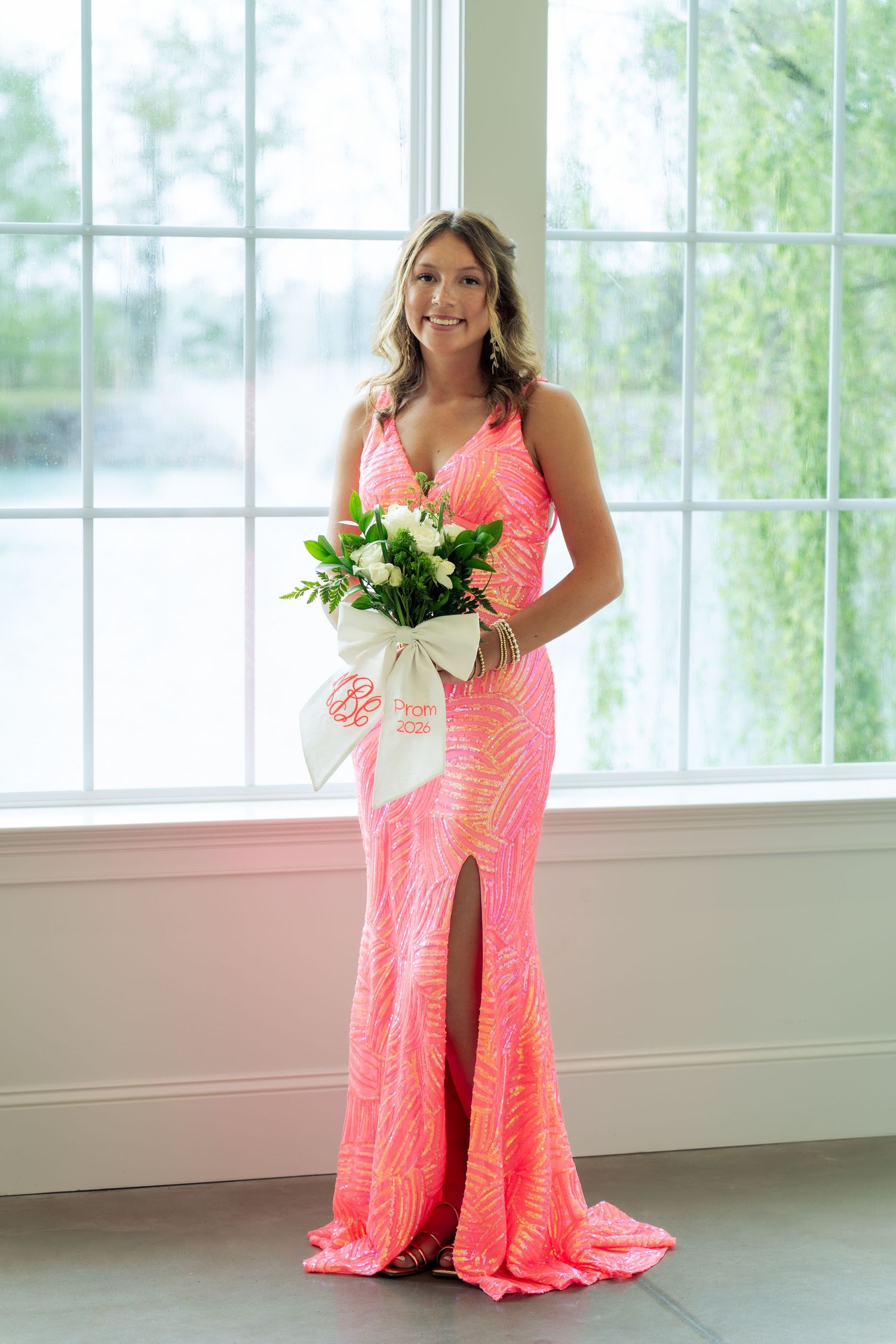 Young woman in a pink sequined gown holding a white bouquet, posing elegantly in front of a large window.