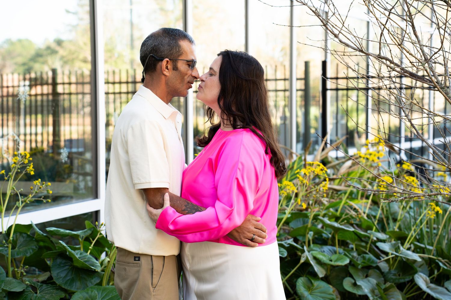 A couple shares a romantic moment outdoors, woman in pink top and man in beige shirt, surrounded by lush greenery.