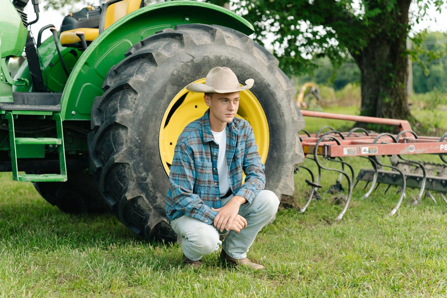Young farmer in cowboy hat crouching by large John Deere tractor tire in green field.