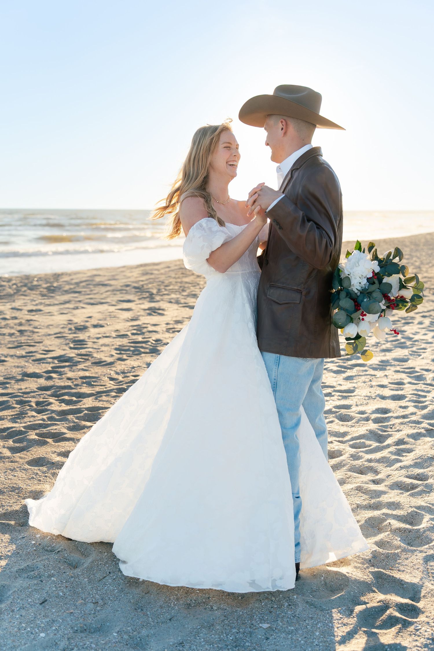 Bride and groom dancing on a sandy beach at sunset, groom holding a bouquet and wearing a cowboy hat.