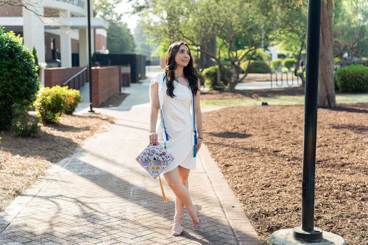 Woman in white dress walking on brick path in sunny park, carrying floral clutch and blue bag.