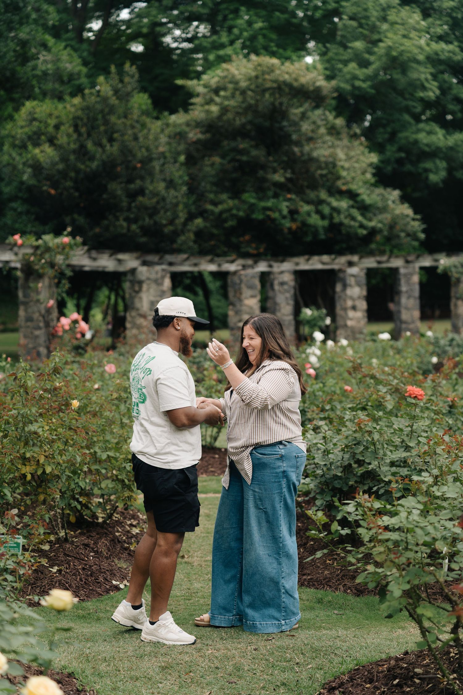 Couple laughing together in a rose garden with stone pergola columns and lush green trees in the background.