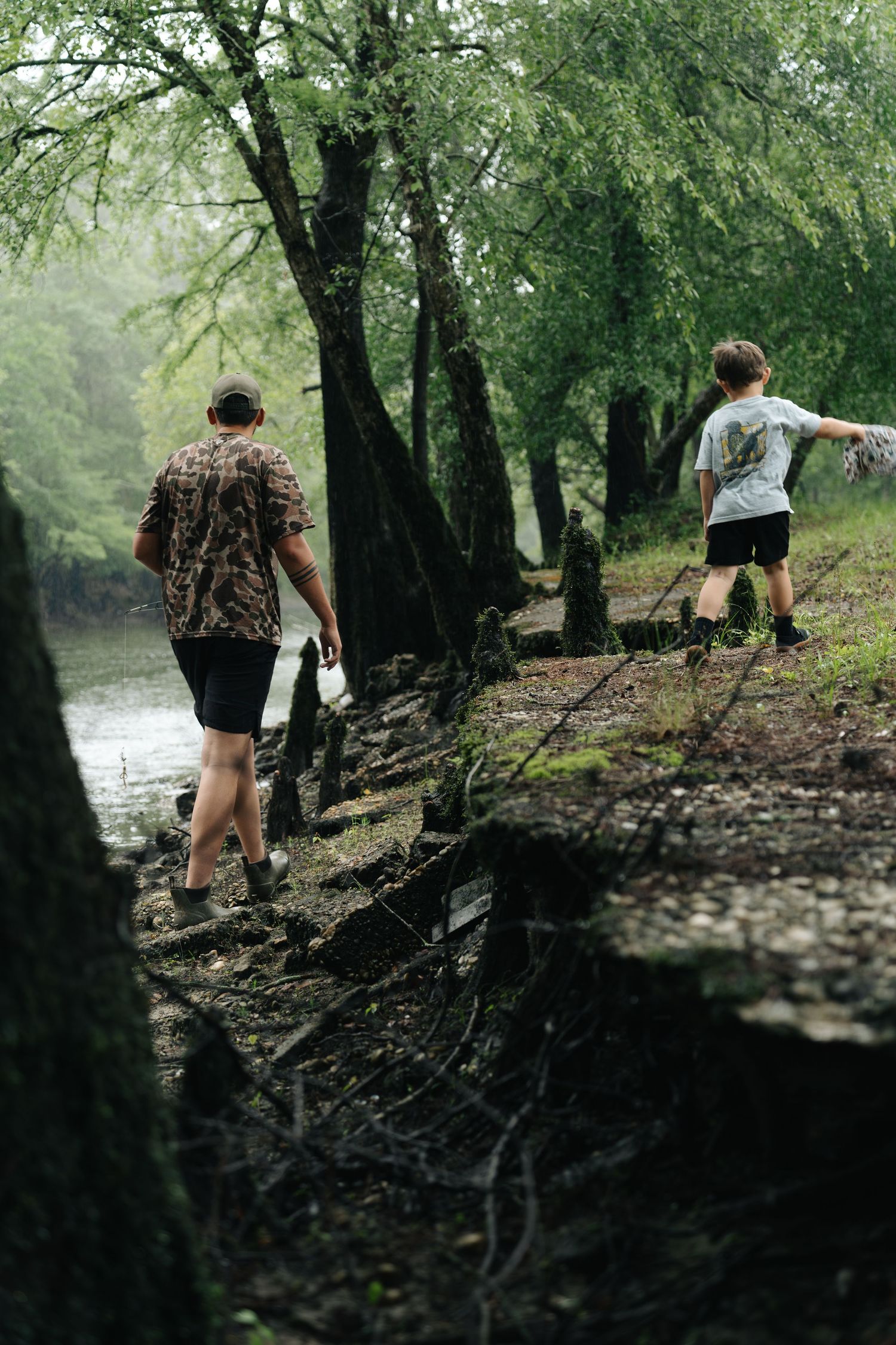 Two people walking along a misty riverbank through a dense forest, with roots and fallen branches on the ground.