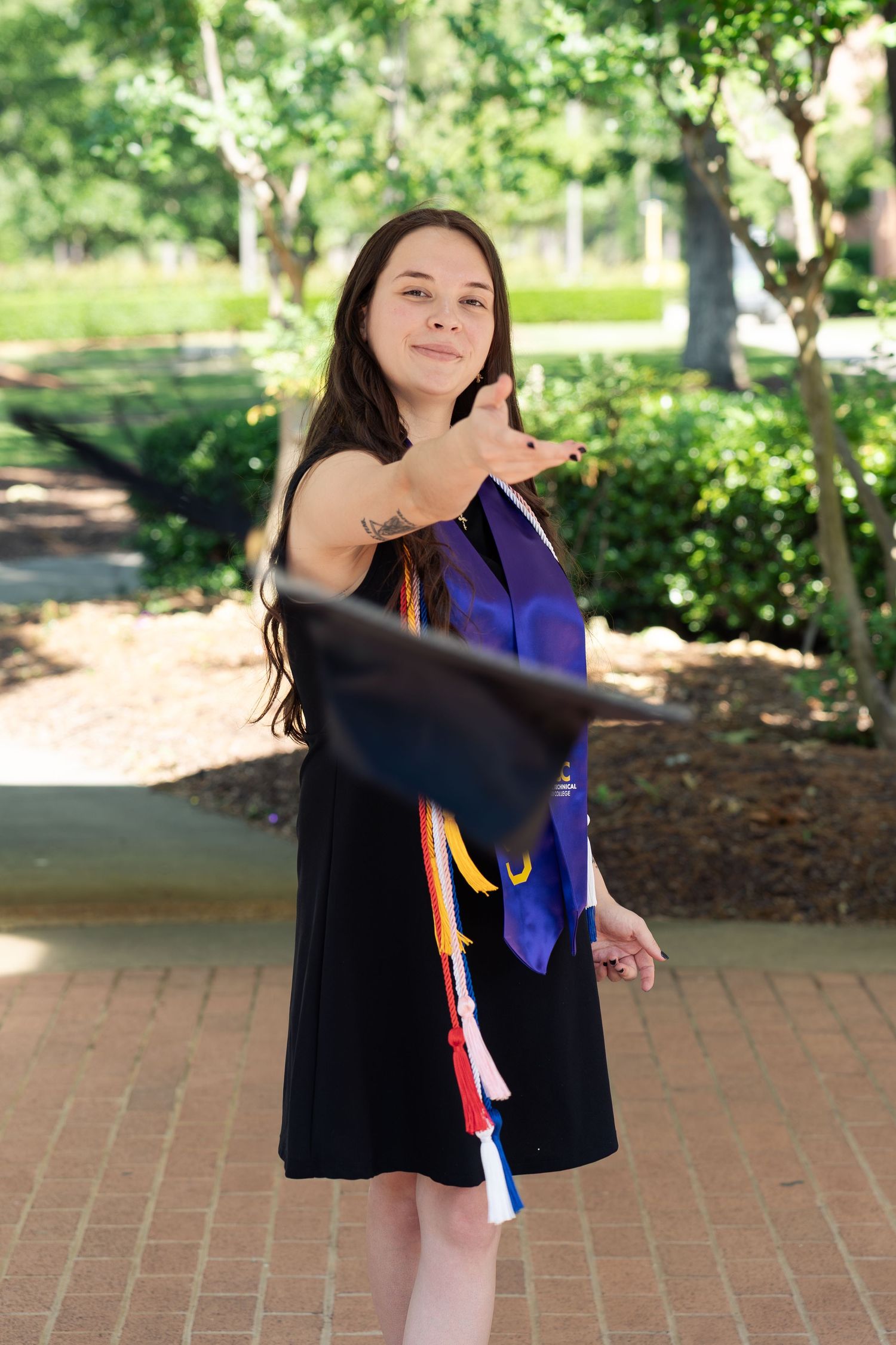 Young woman in graduation attire tossing cap outdoors on campus walkway surrounded by lush green trees.