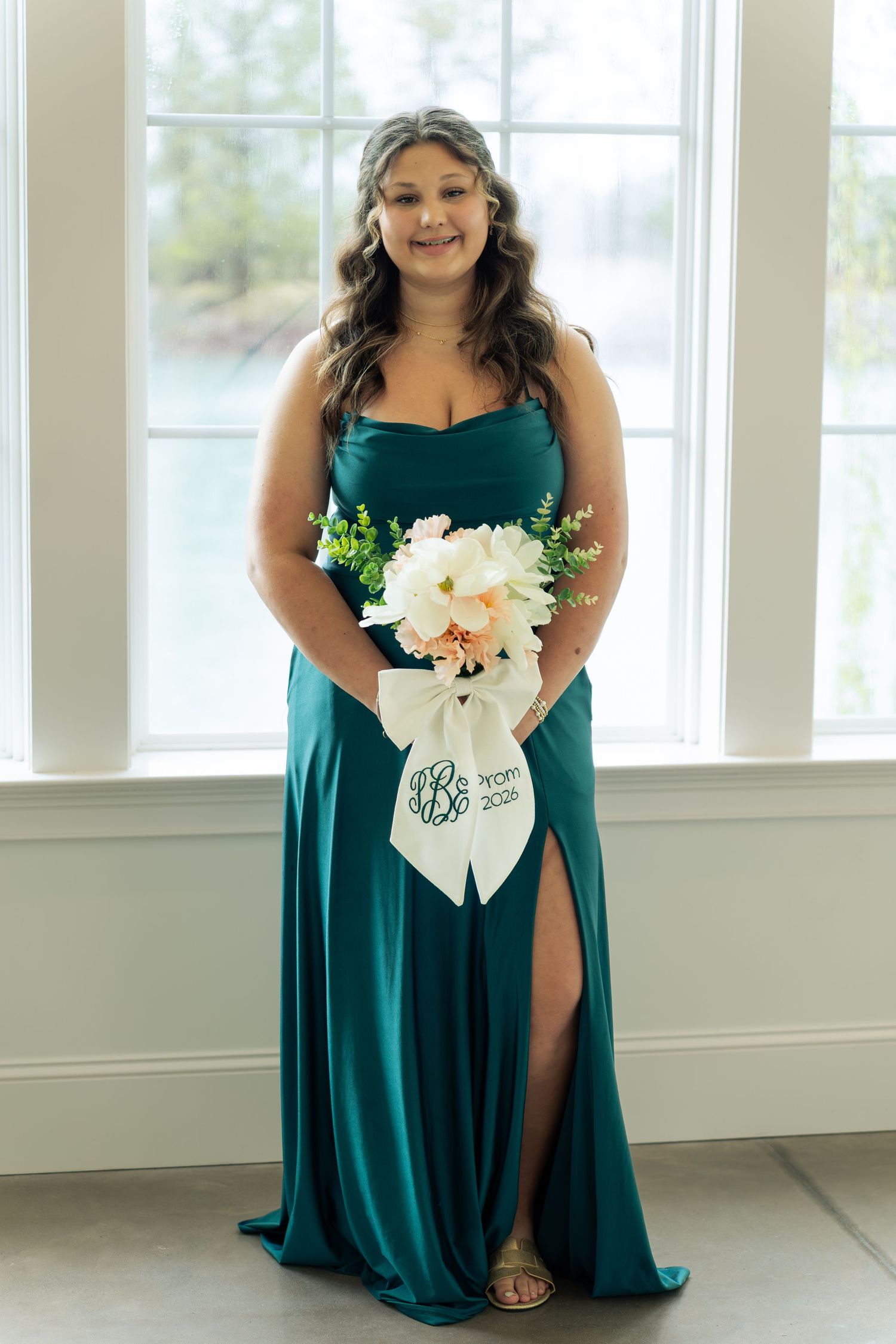 Smiling bridesmaid in emerald green gown holding white floral bouquet with monogram ribbon stands by window.