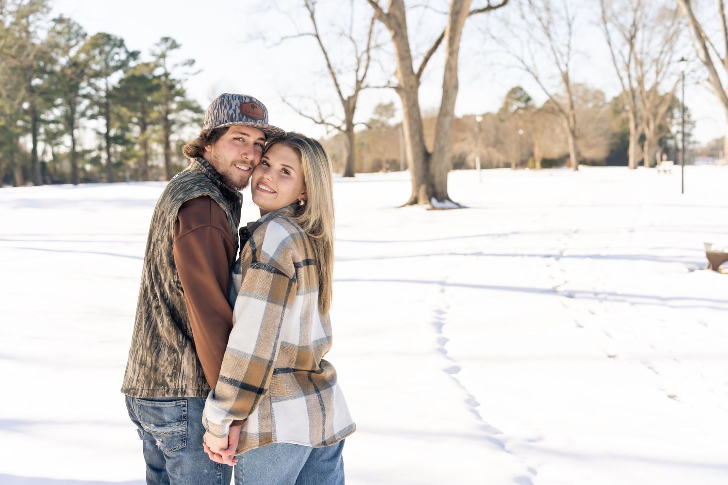 Couple embracing and smiling in a snowy winter park surrounded by bare trees on a sunny day.