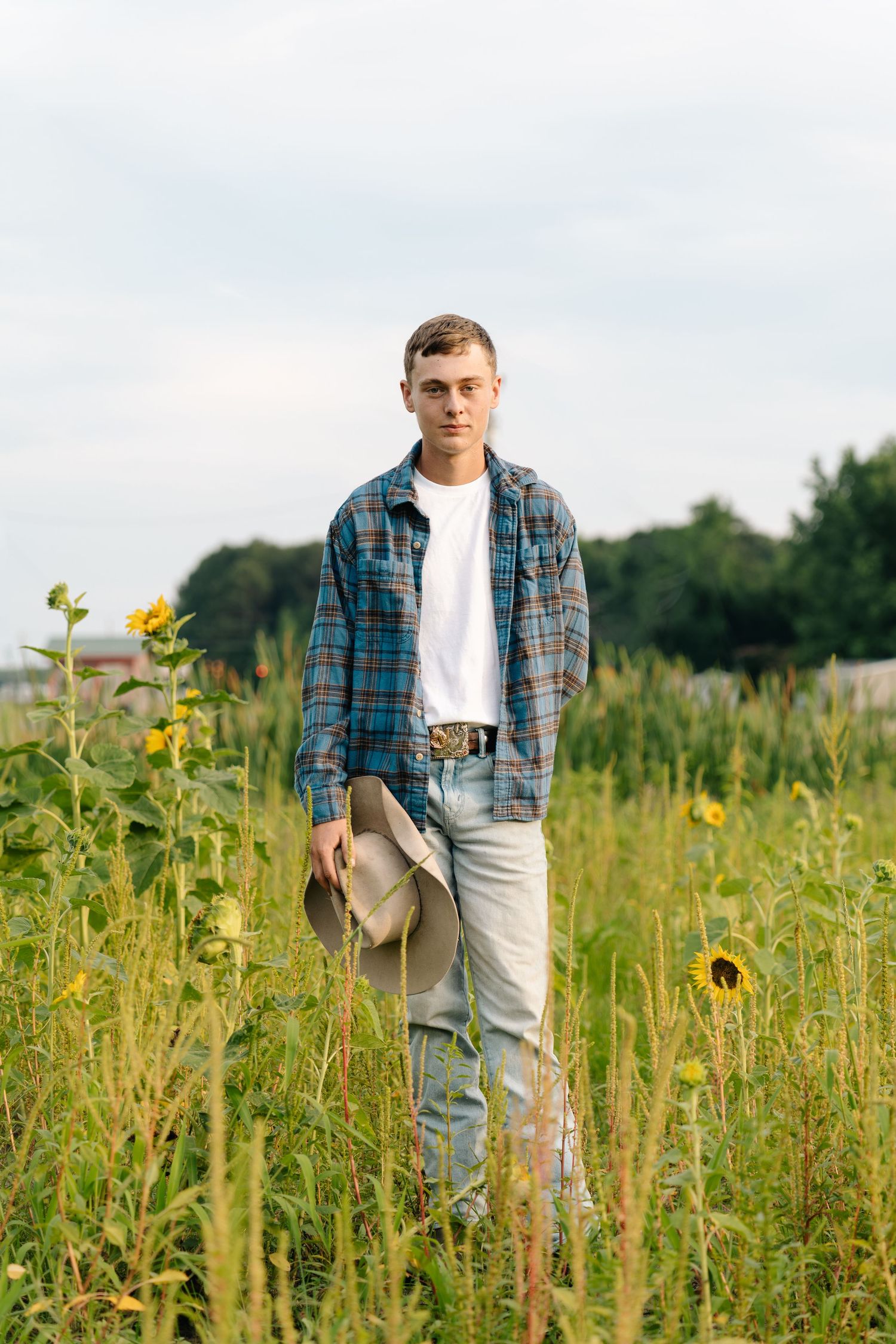 Young man in plaid shirt and jeans holding cowboy hat standing among sunflowers in a rural field.