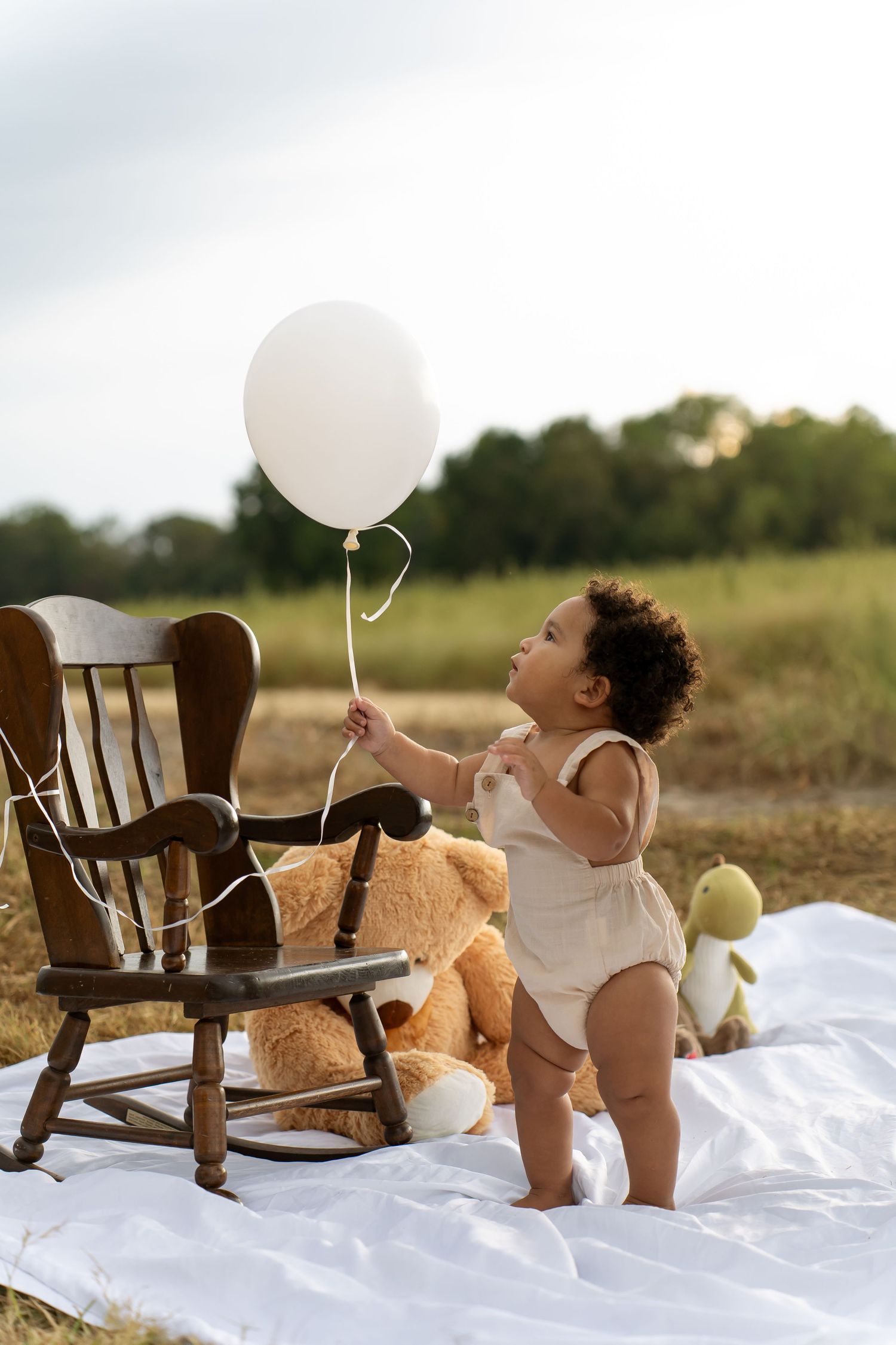 Baby playing with white balloons and teddy bear on white blanket in outdoor setting.