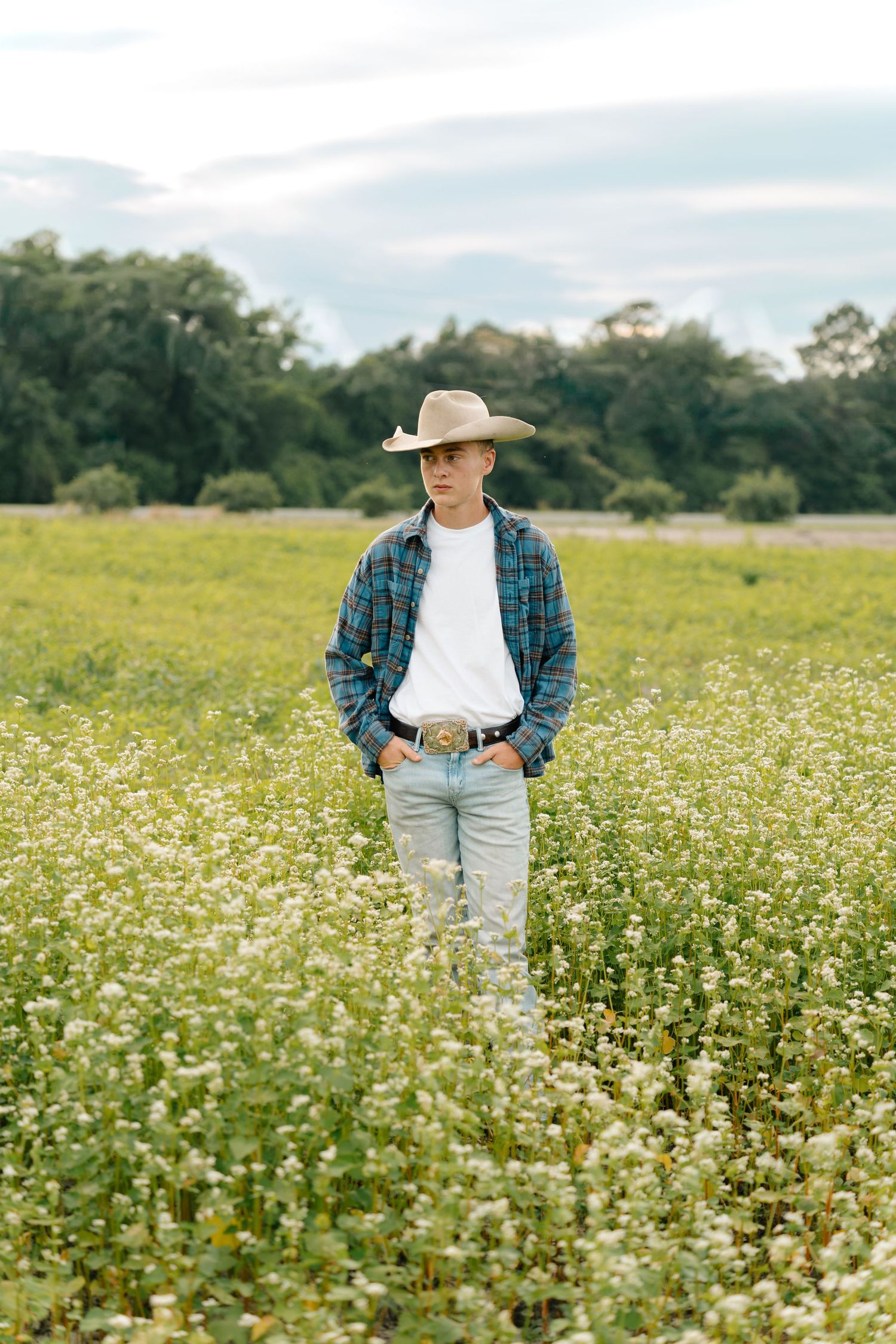 Young man in cowboy hat, plaid shirt, and jeans standing in a blooming wildflower field with trees in background.