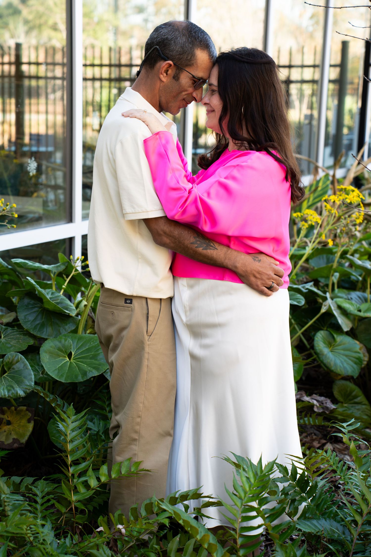A couple embraces tenderly among lush green plants, woman in pink top and white skirt, man in beige outfit.