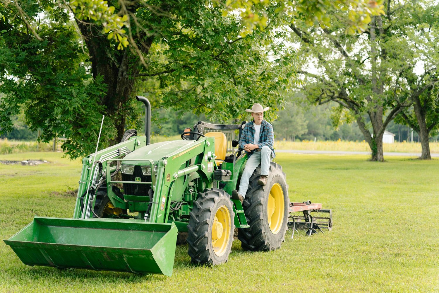 Farmer driving a green John Deere tractor with front loader on a lush farm property under large shade trees.