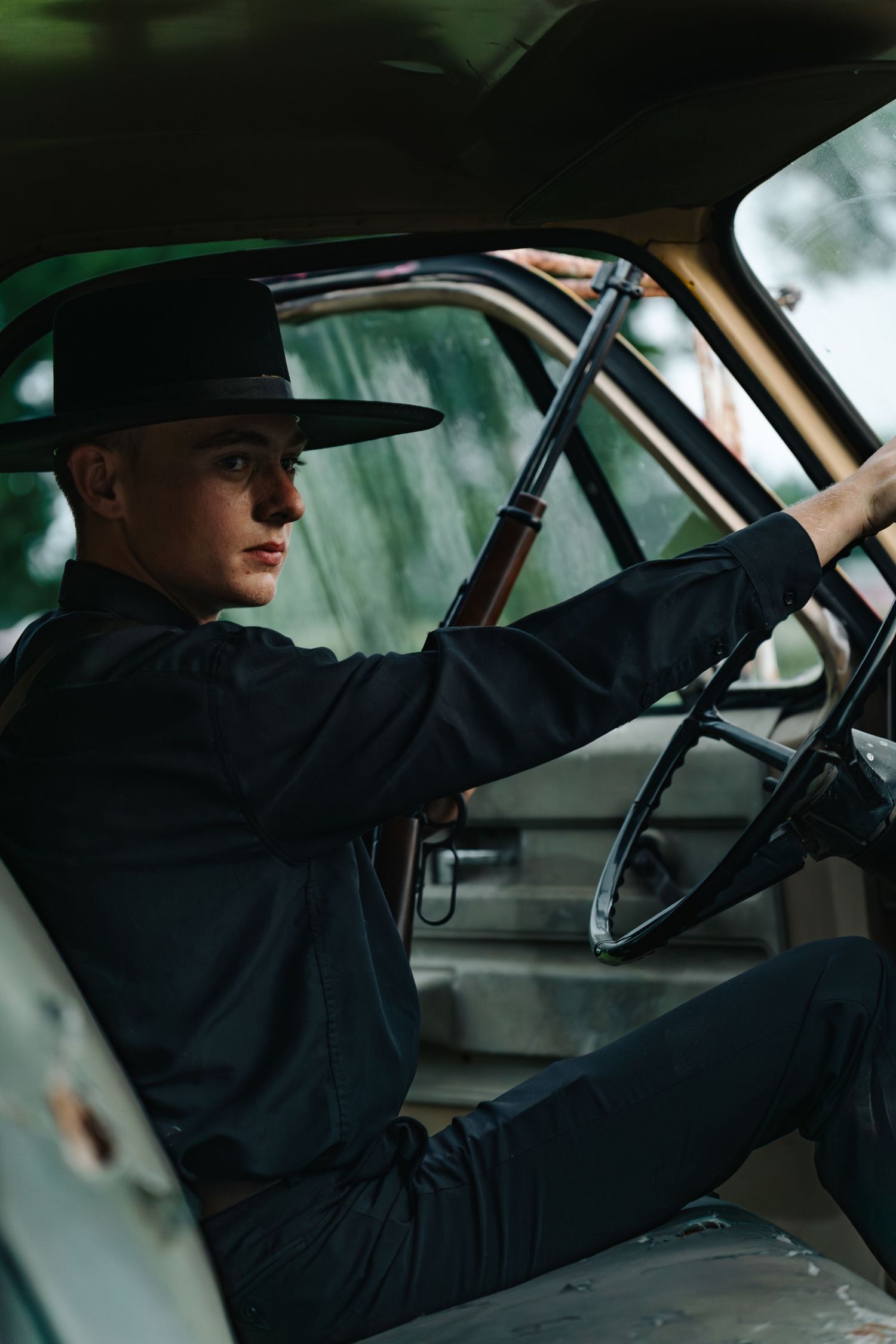 Young man in black outfit and wide-brim hat sitting in vintage vehicle holding a rifle.