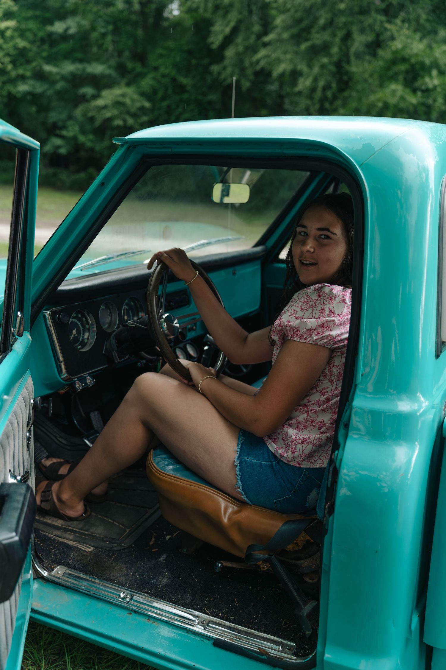 Young woman in denim shorts sitting in vintage turquoise pickup truck cab, smiling at camera while holding steering wheel.