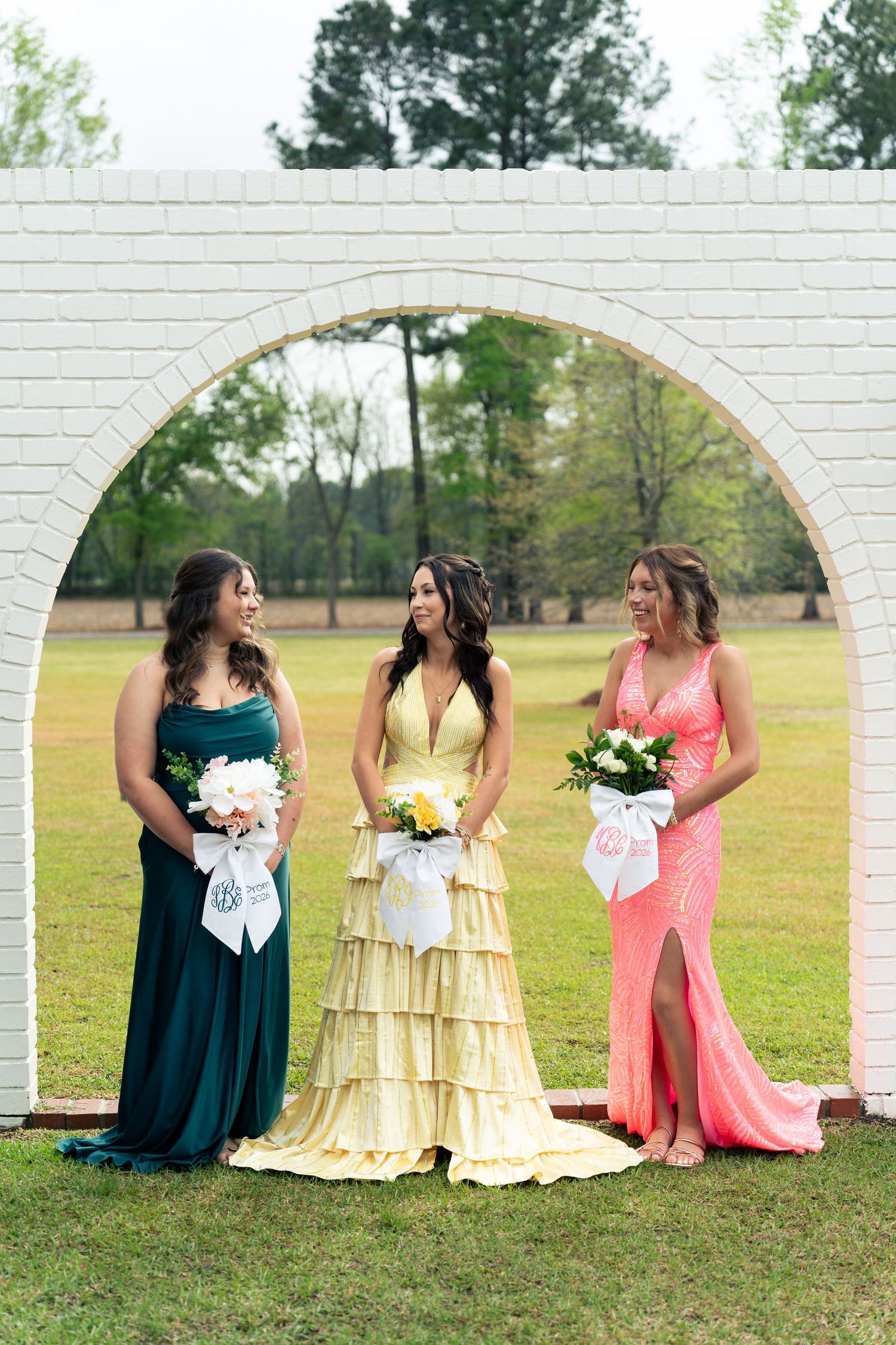 Bride in yellow ruffled gown stands with two bridesmaids in teal and coral dresses under white brick arch.