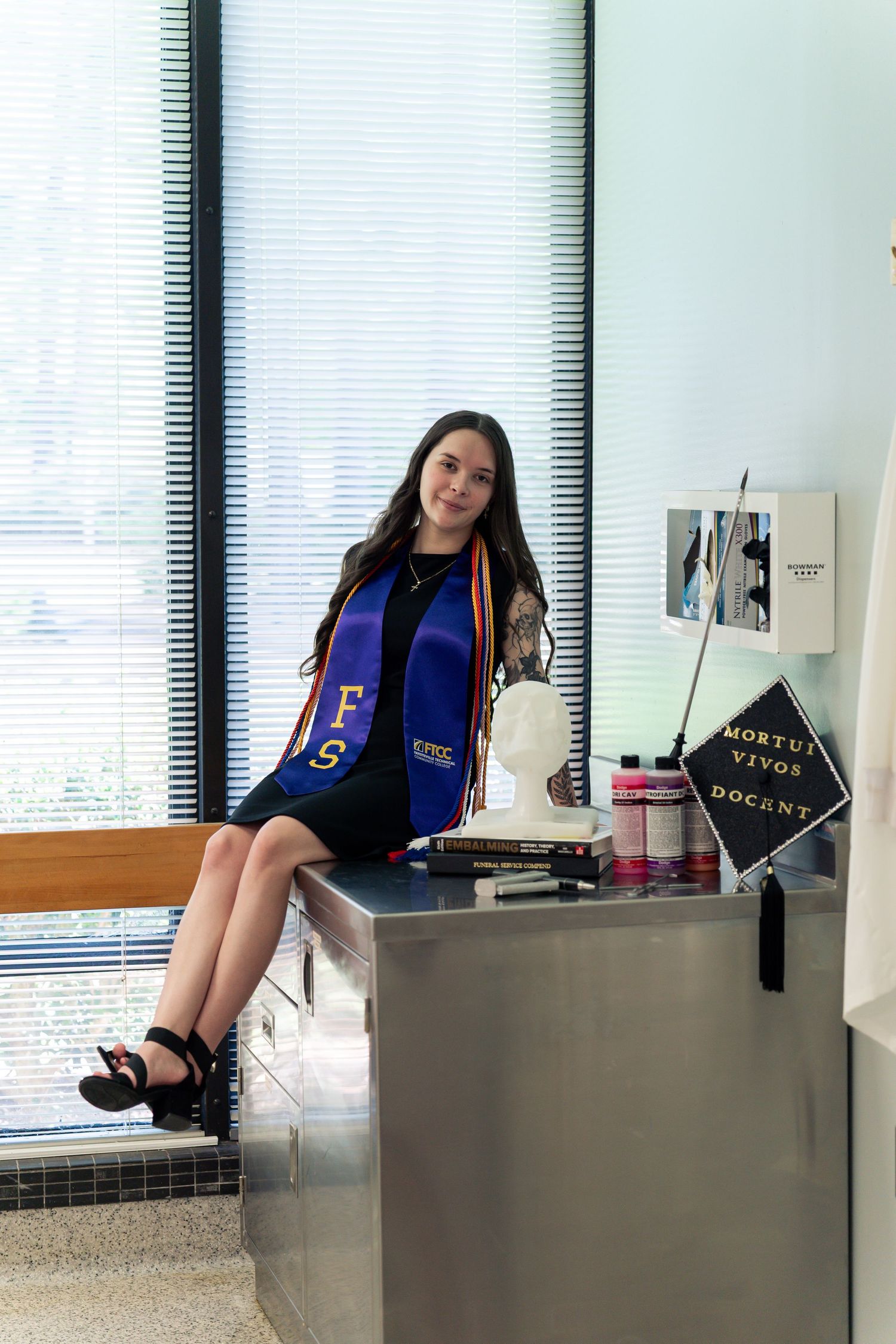 Graduate in blue sash sits on metal counter near mortuary sign, celebrating academic achievement.