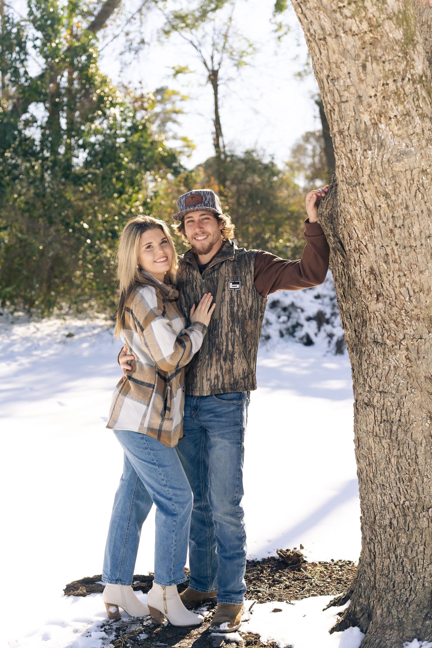A smiling couple poses together by a tree in a snowy winter landscape, wearing casual plaid and camo outfits.