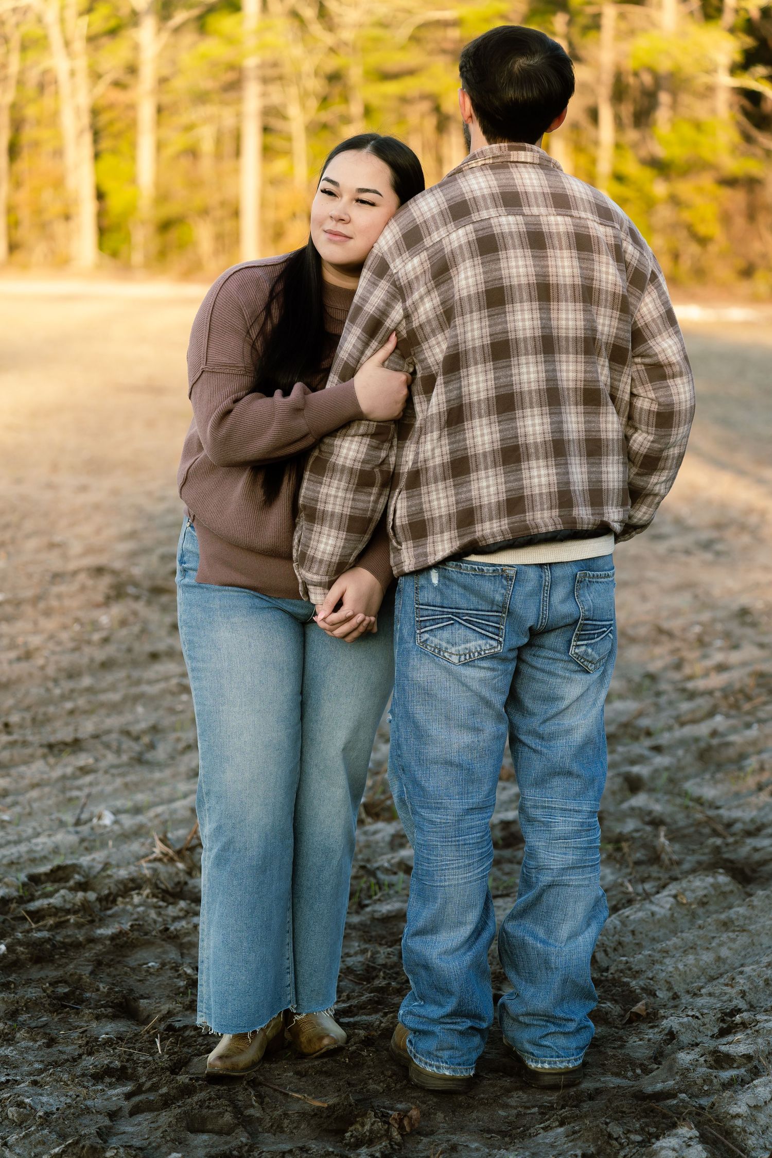 A couple embraces in a muddy field, woman in brown sweater and jeans leaning on man in plaid shirt at golden hour.