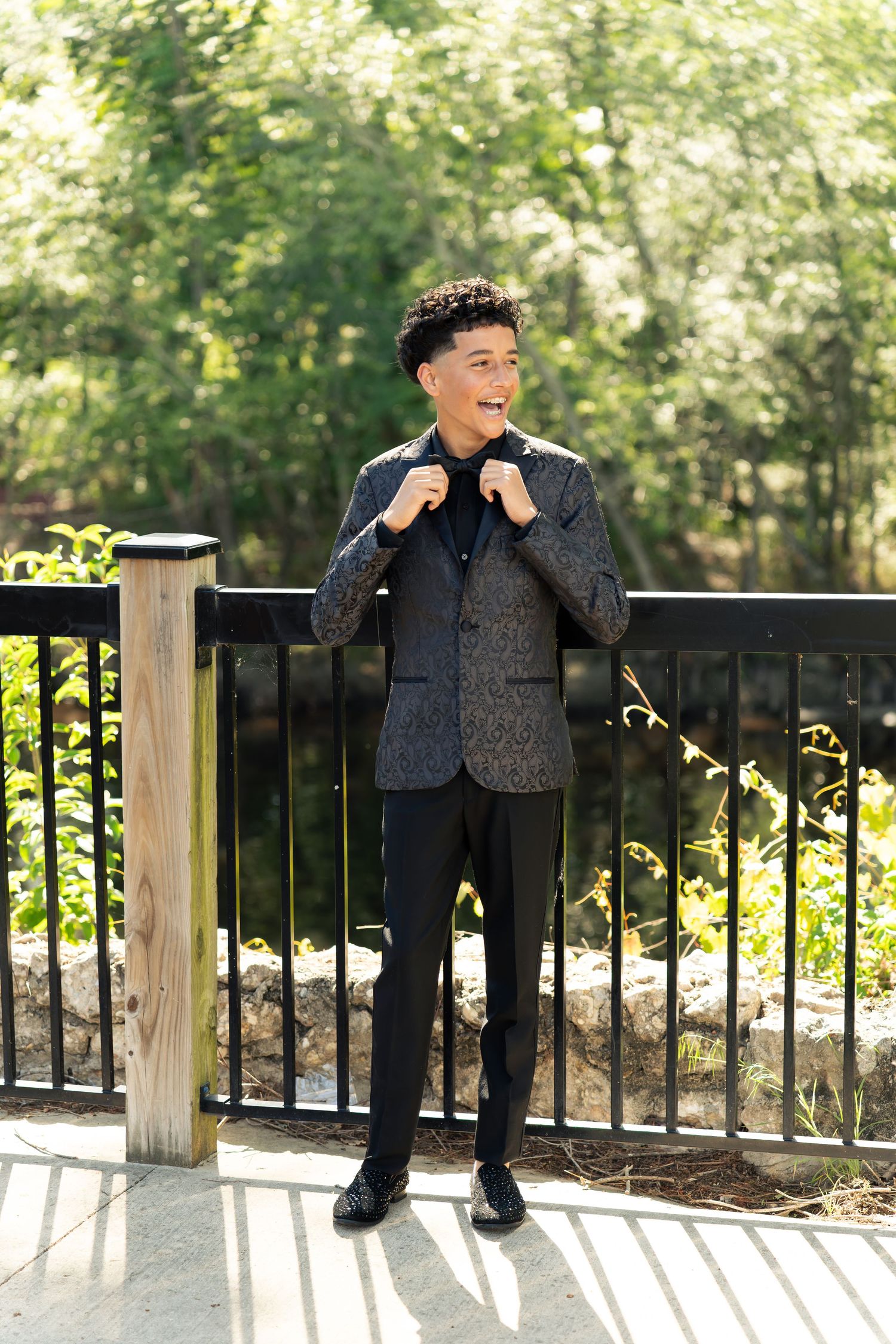 Young man in patterned blazer and black pants posing outdoors near metal railing with green trees background.