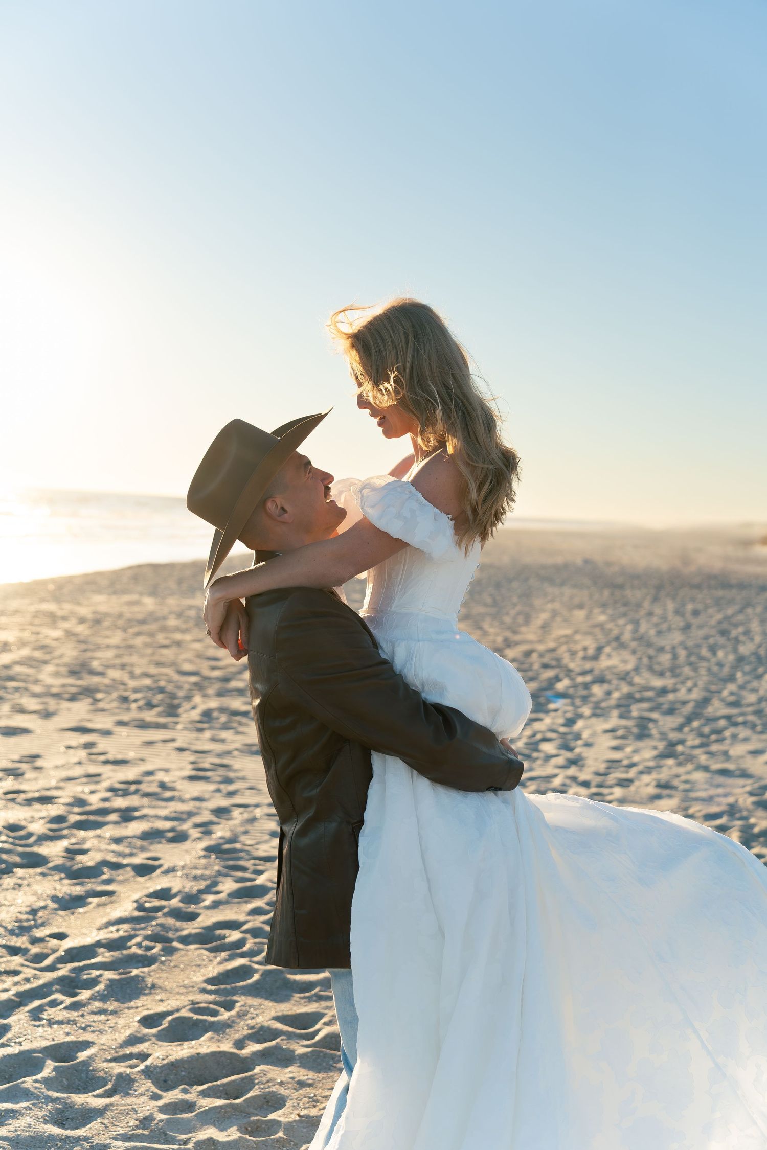 Groom lifting bride in white dress on sunny beach, romantic wedding photo with golden sand and clear sky.