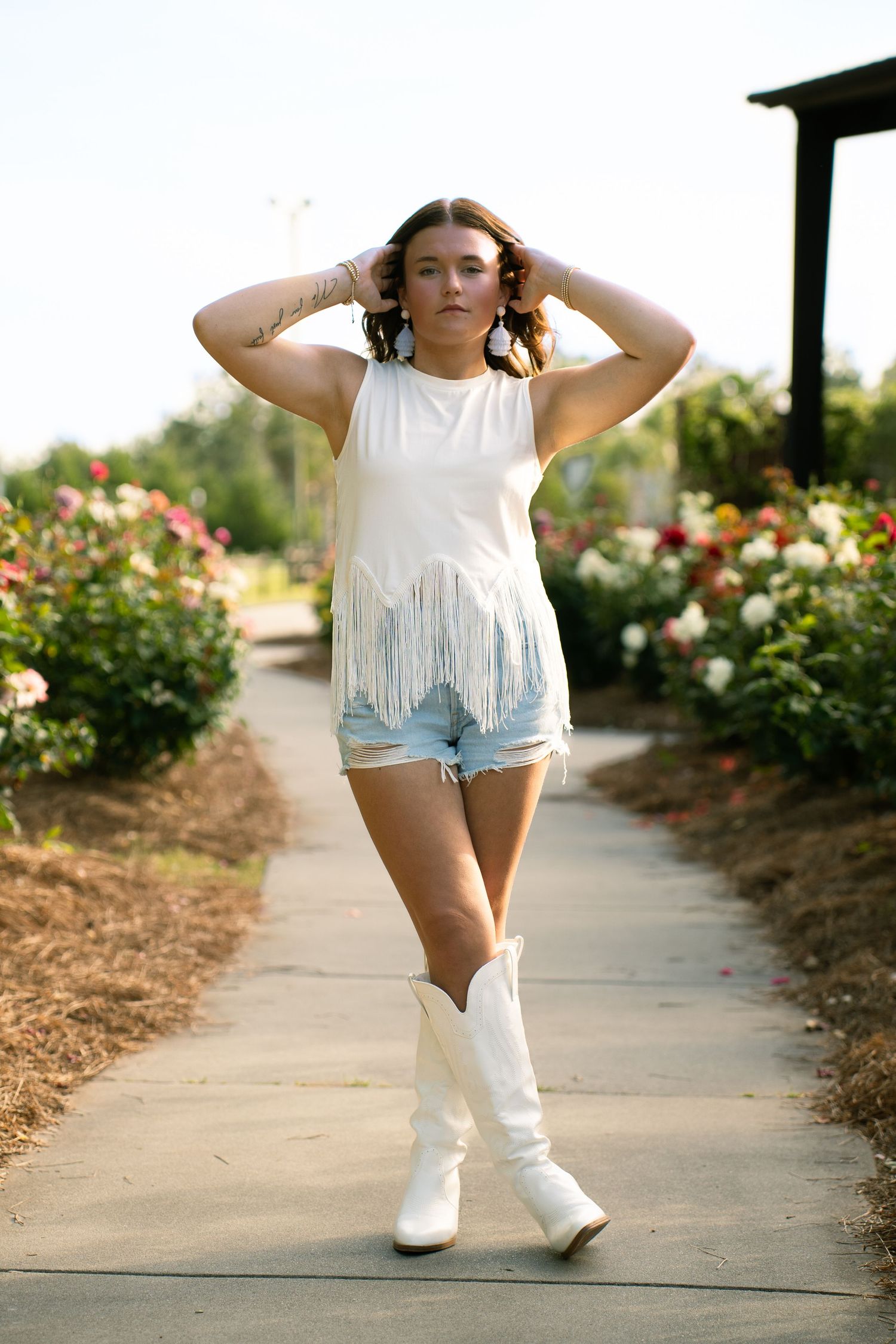 Person in white fringe top and boots poses on walkway near blooming rose bushes.