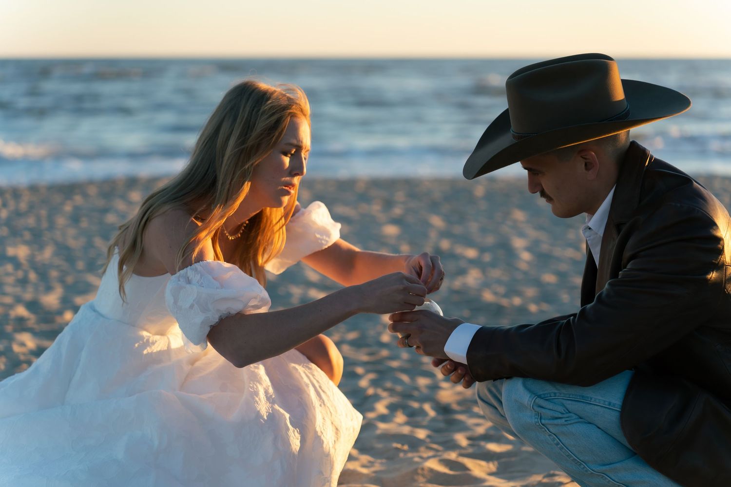 Couple in wedding attire sharing a romantic moment on a beach at sunset, groom in cowboy hat holding bride's hands.