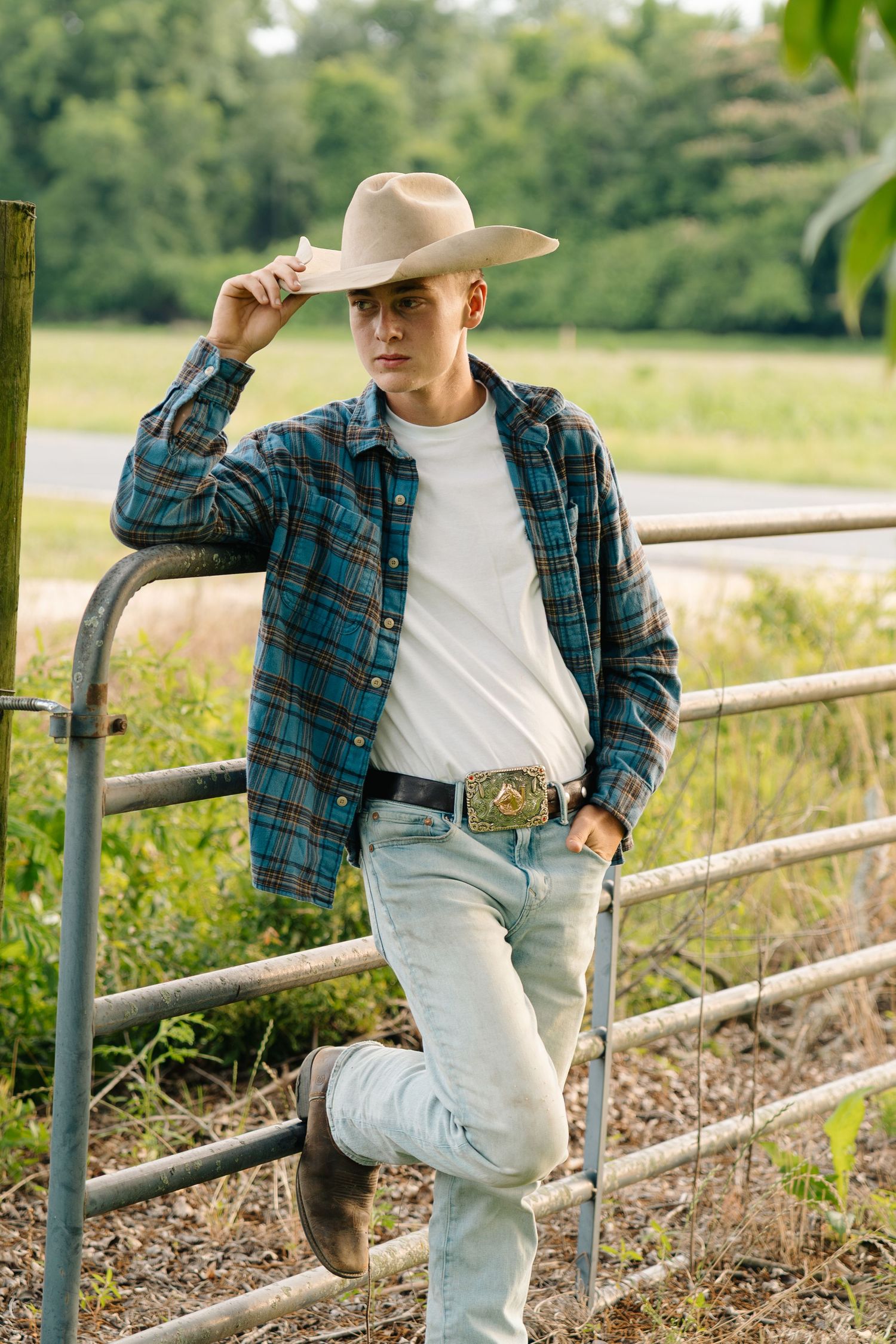 Young man in cowboy hat, plaid shirt, and jeans leaning on a farm gate in a rural countryside setting.