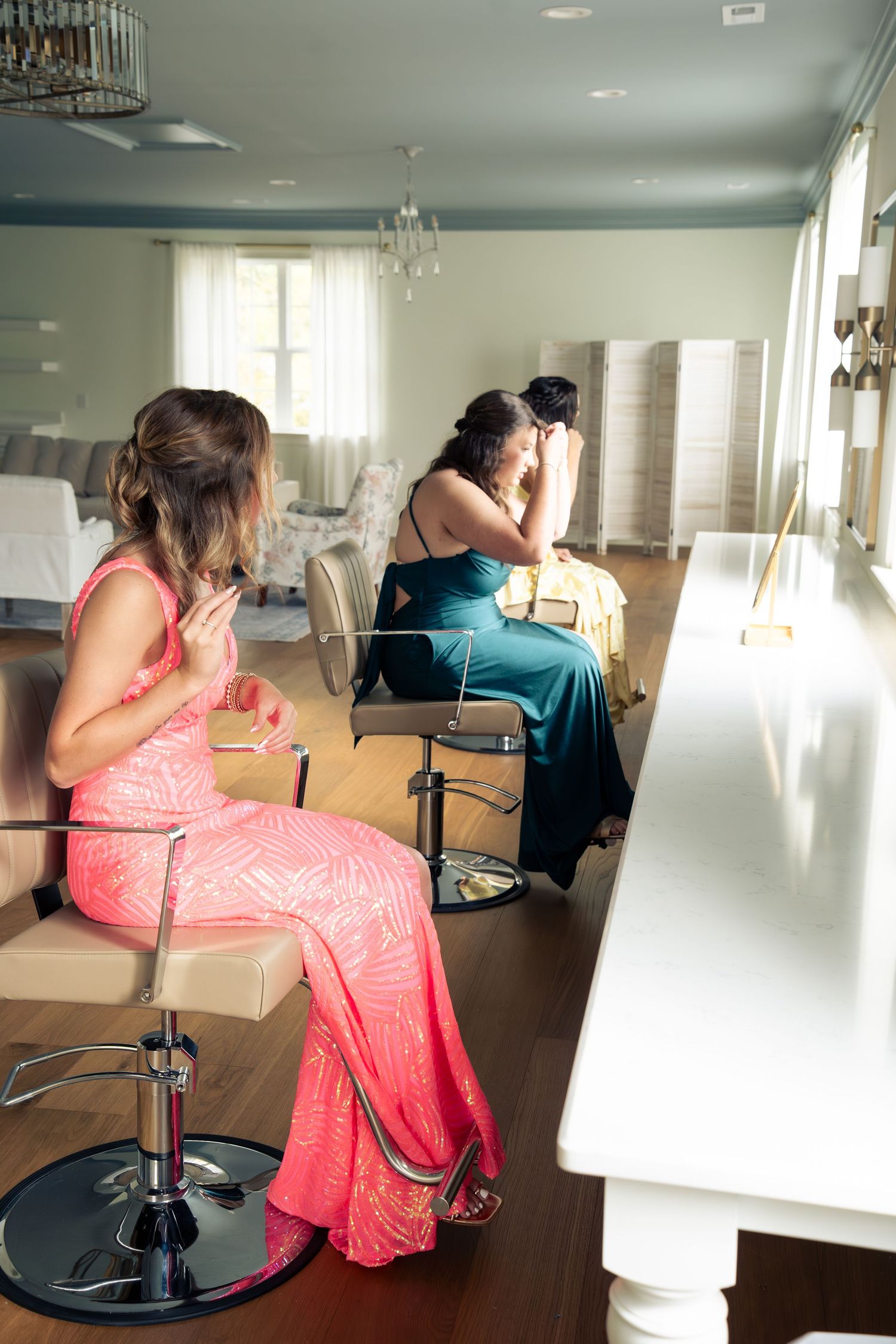 Two bridesmaids in pink and teal gowns sit in salon chairs preparing for a wedding ceremony.