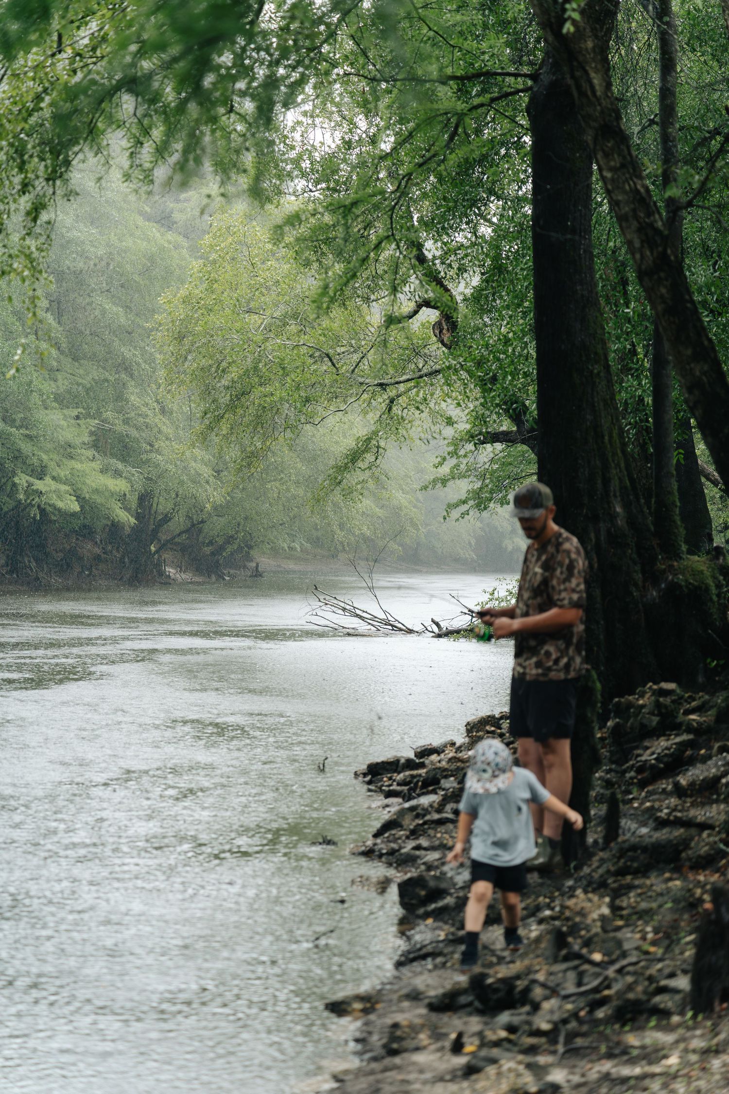 Adult and child fishing on a misty river bank surrounded by lush green trees in a serene forest setting.