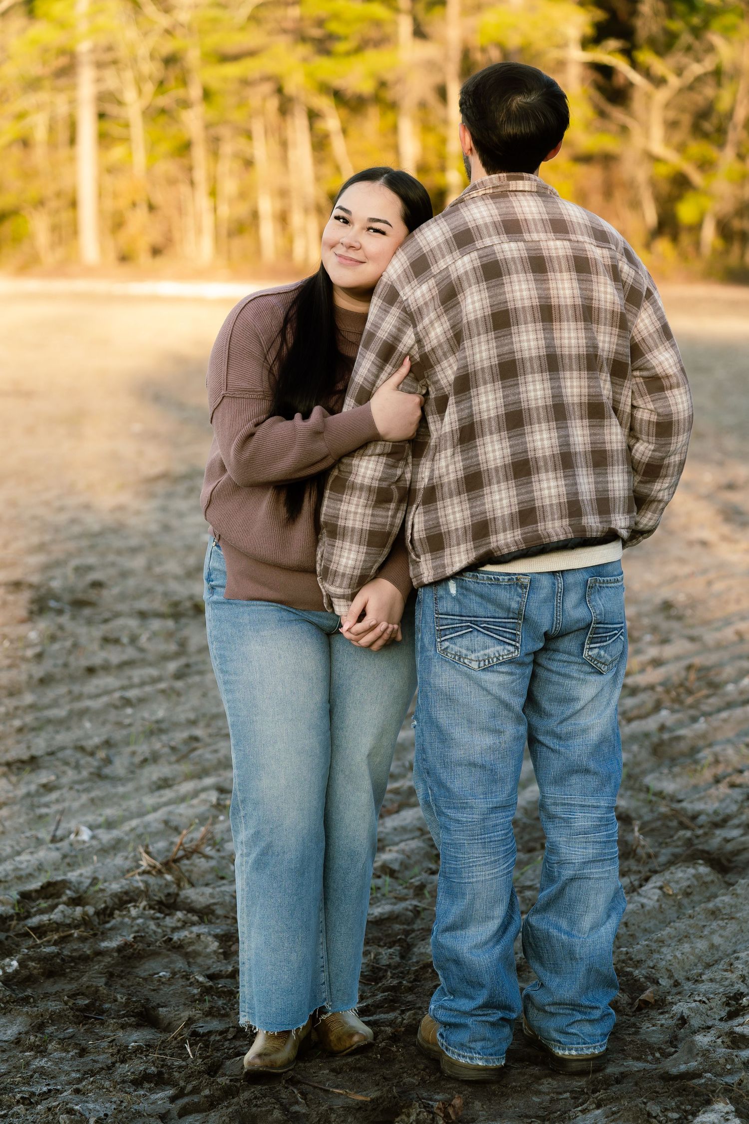 Couple embracing outdoors in casual fall attire, woman smiling over shoulder, trees in golden light background.