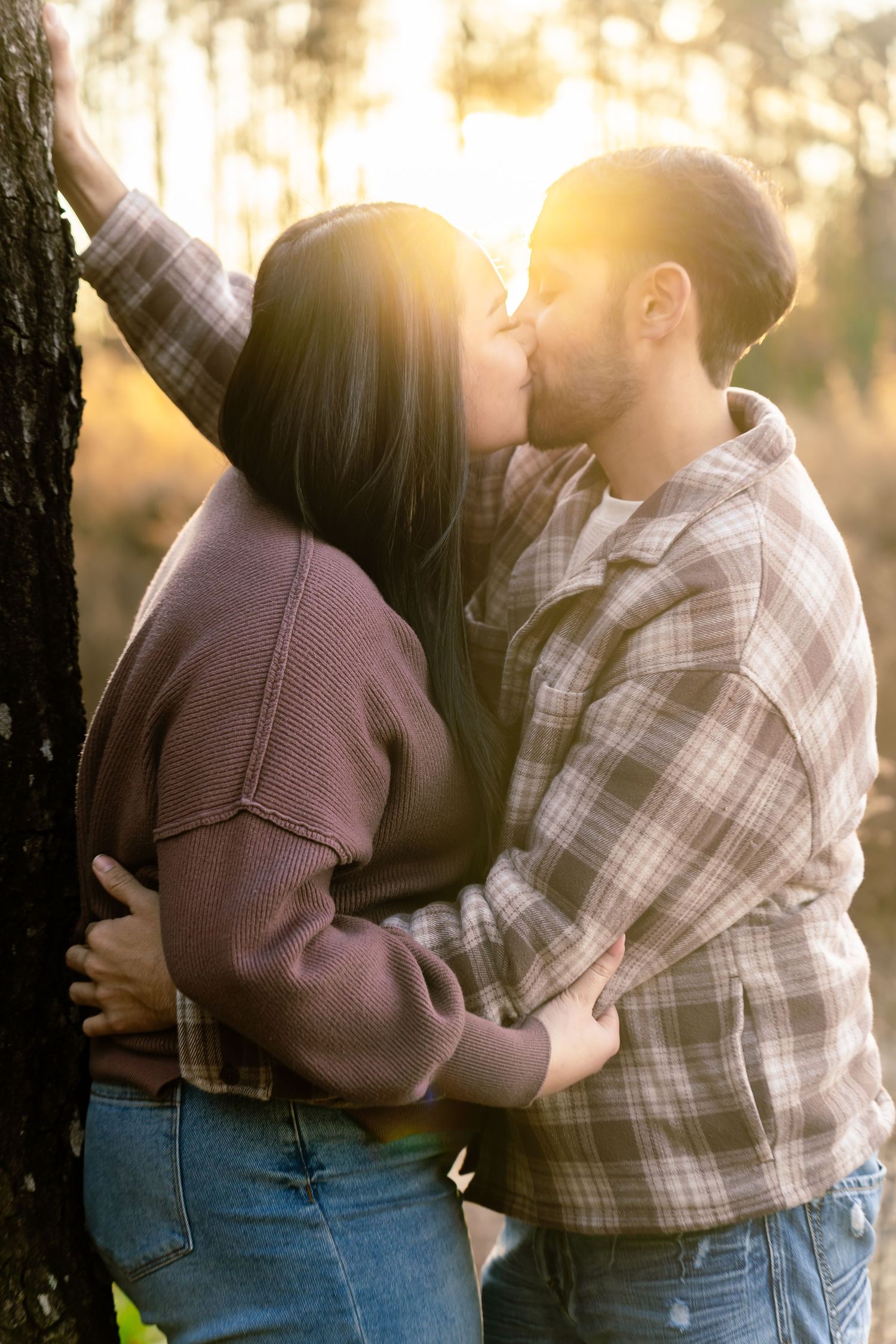 Couple sharing a romantic kiss outdoors near a tree, bathed in warm golden sunset light in a forest setting.
