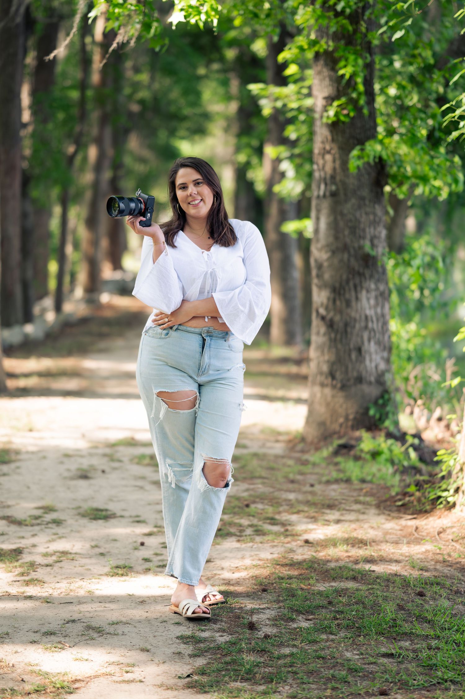 Woman photographer holding camera in ripped jeans and white top on a tree-lined path outdoors.