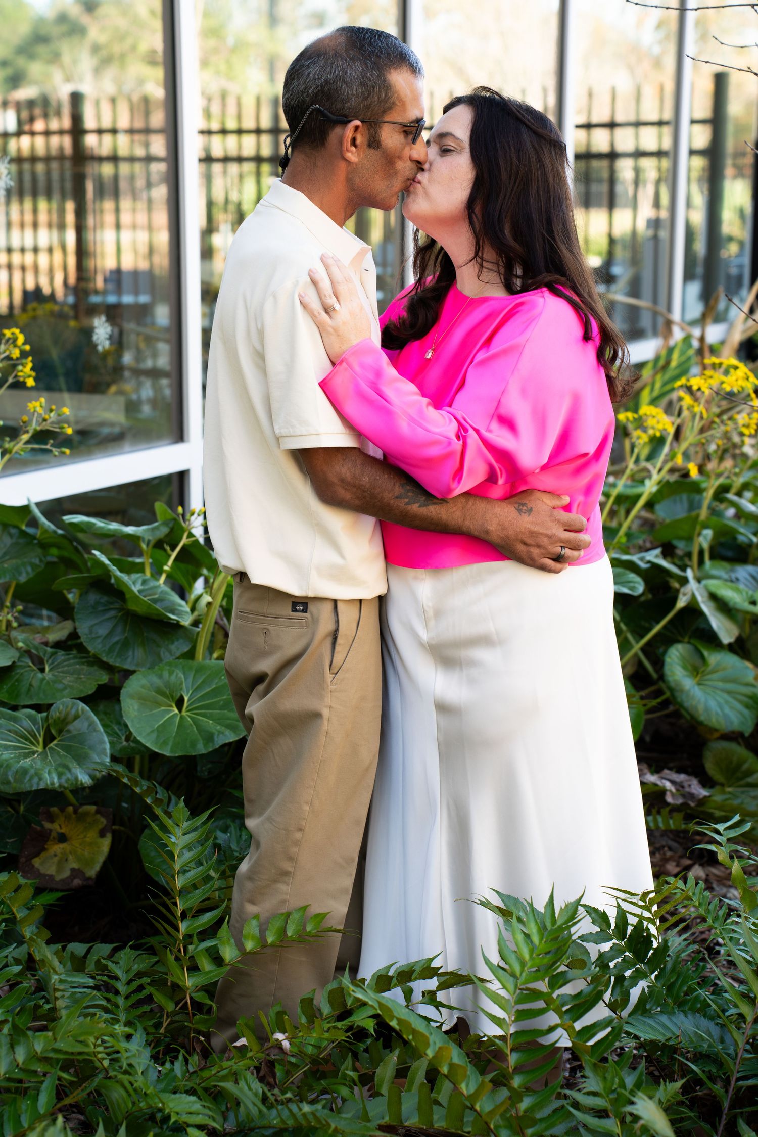 A couple shares a romantic kiss in a garden setting with lush greenery and a black iron fence in the background.