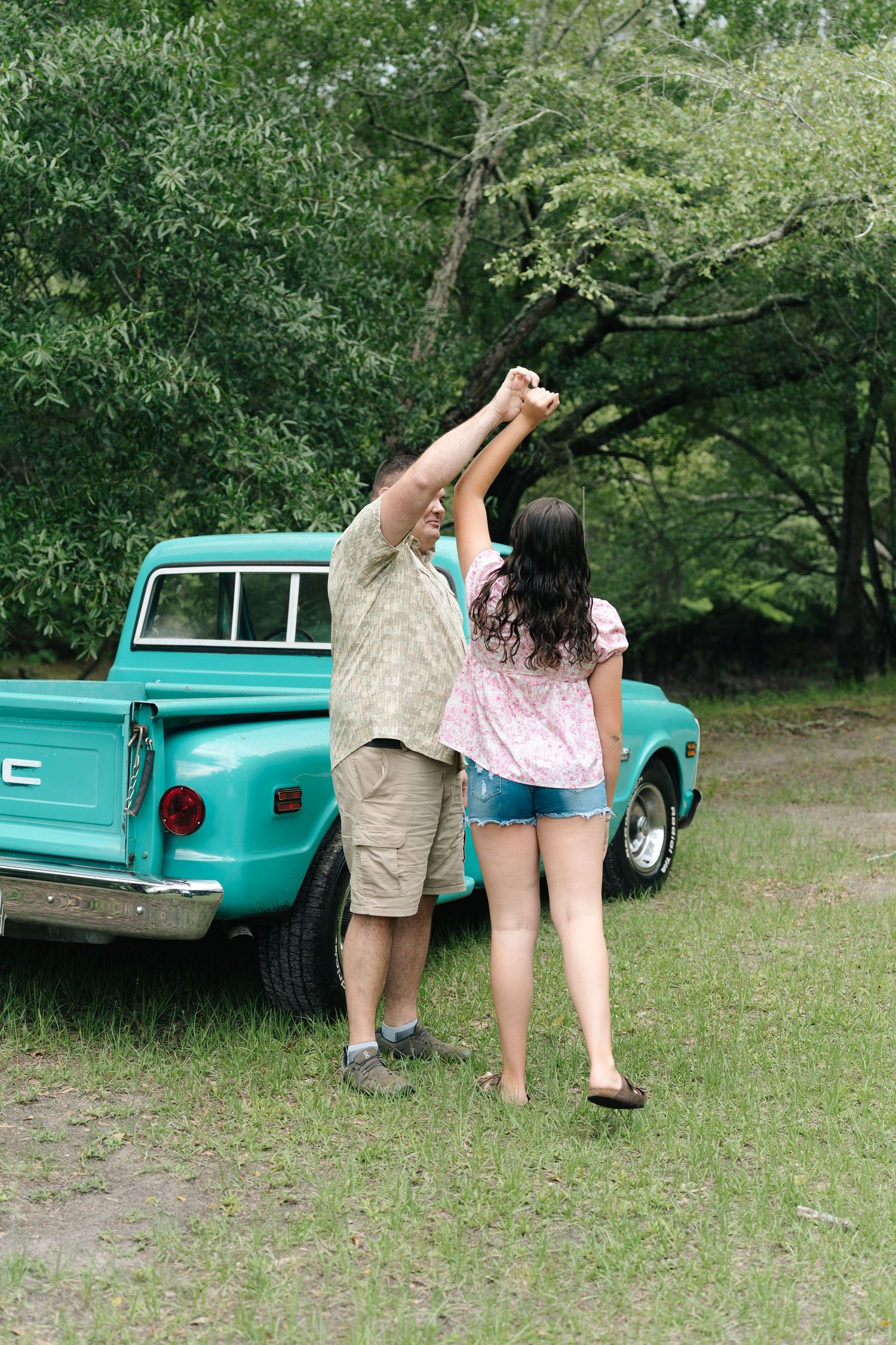 A couple holds hands raised while standing behind a classic teal pickup truck in a lush green setting.