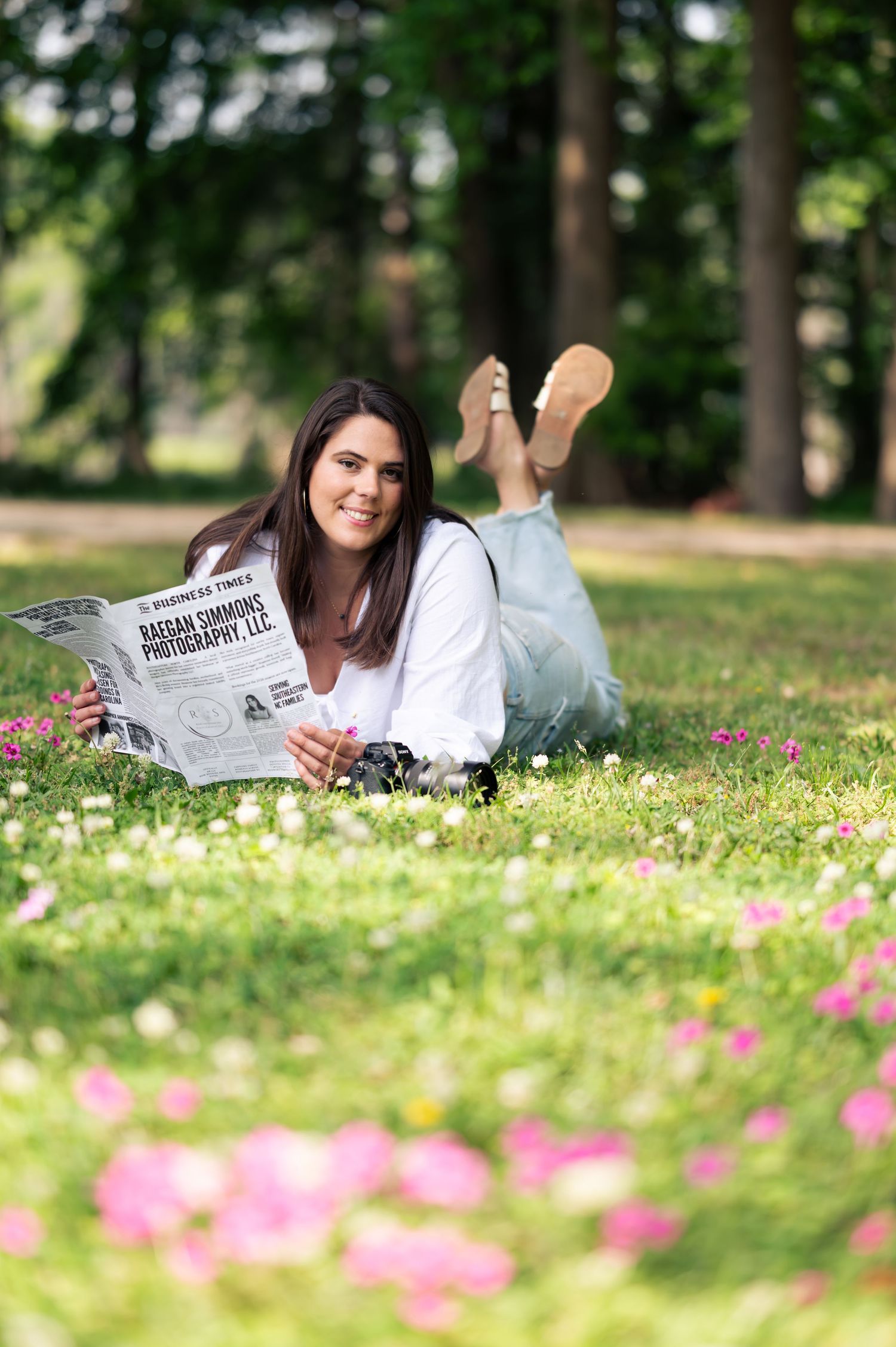 Woman lying on grass in a flower-filled park, smiling and reading a Raigan Simmons Photography newspaper.