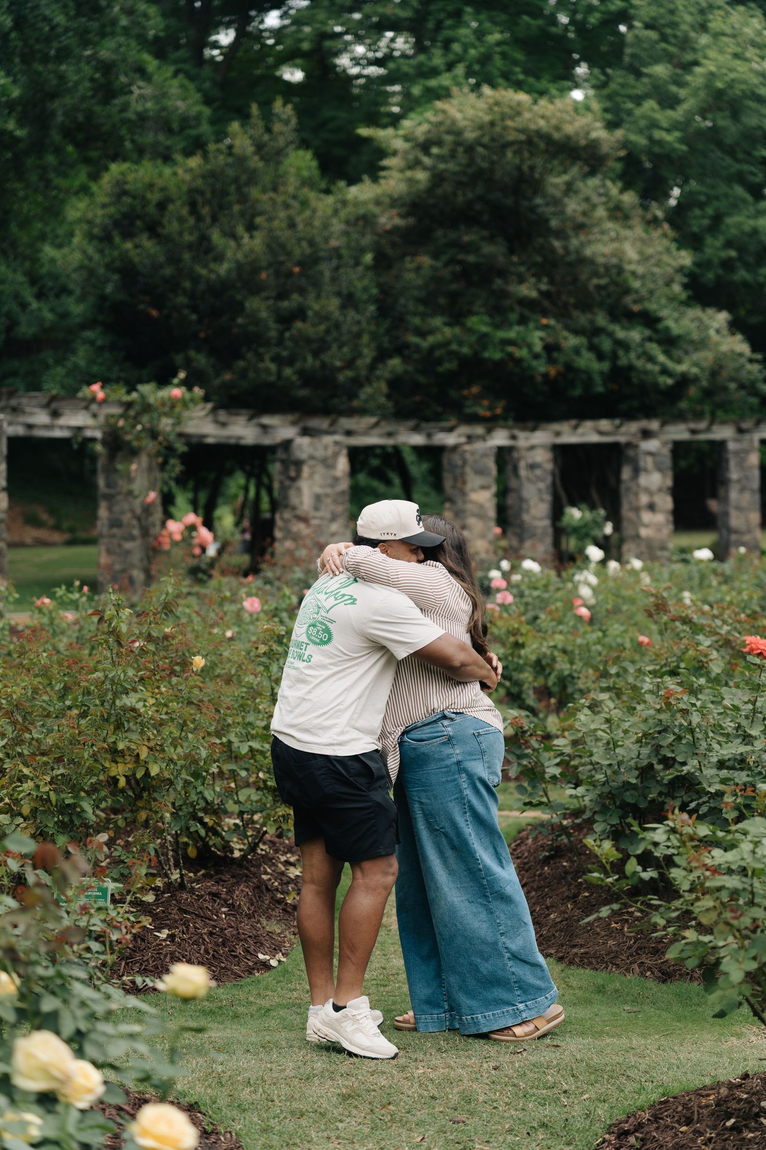 Two people share a warm embrace in a colorful rose garden with stone pergola backdrop.