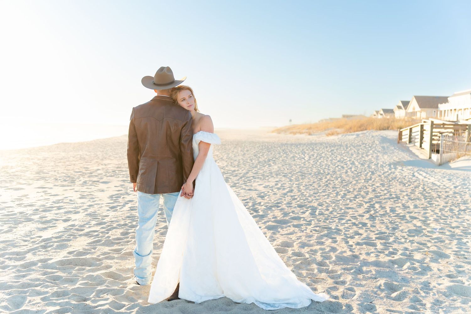 Bride and groom walk hand-in-hand on a sunny beach, she in a white gown, he in a brown suit and cowboy hat.