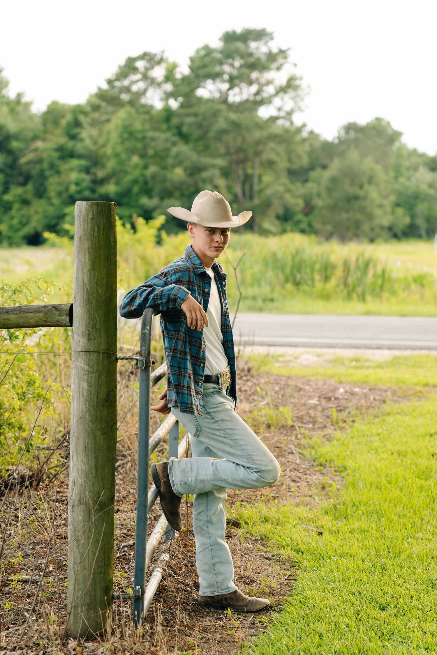 Young man in plaid shirt and cowboy hat leaning against wooden fence post on a rural farm.