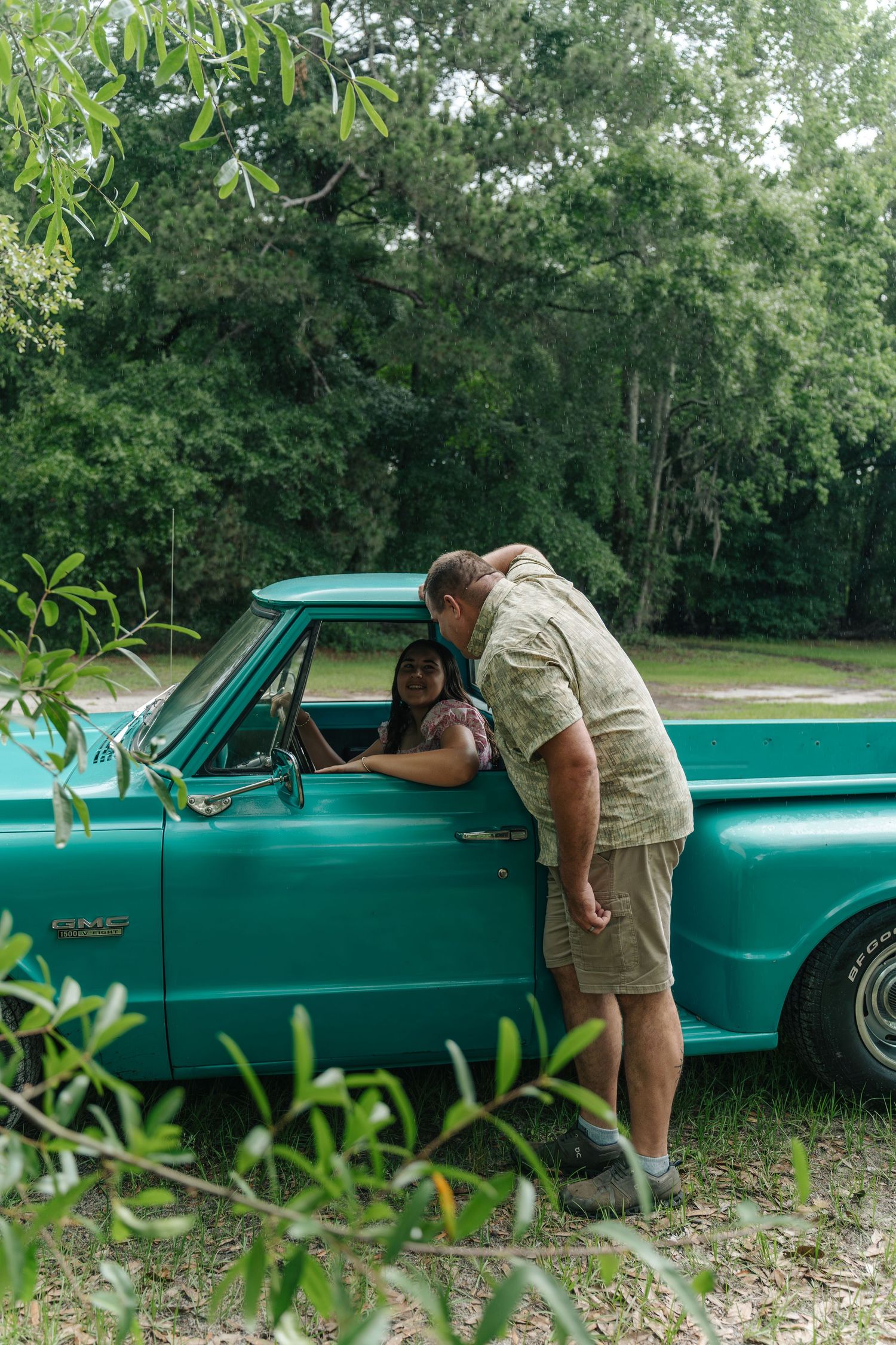 A man leans into a vintage teal GMC truck window talking to a woman inside, surrounded by lush green trees.