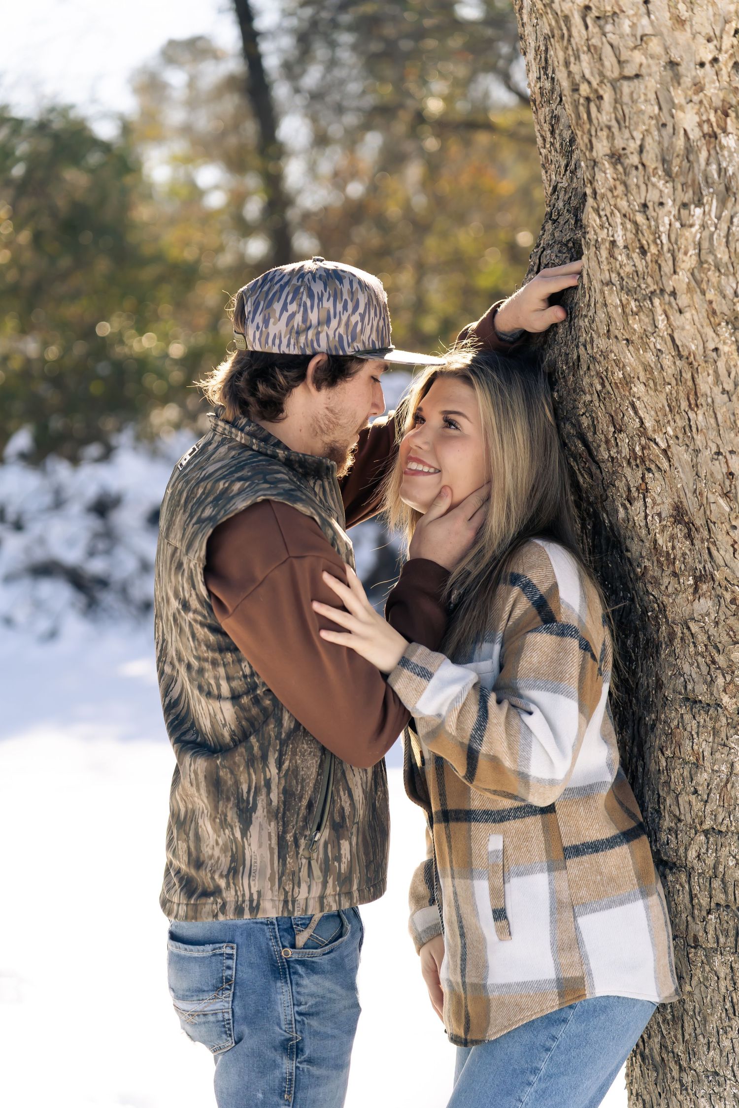 Young couple sharing a romantic moment leaning against a tree trunk in a snowy winter park.