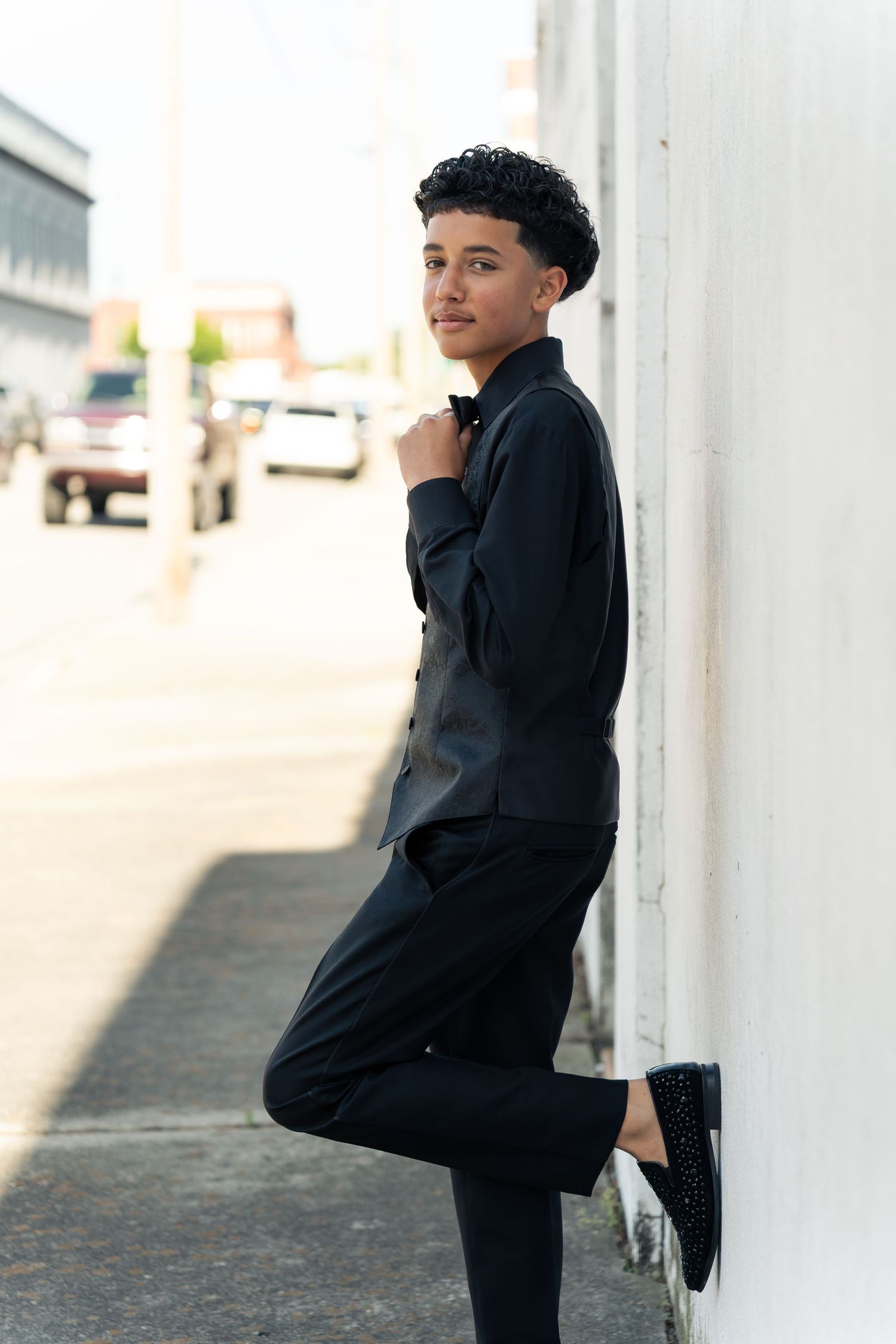 Young person in all-black outfit leaning against white wall on urban street, one leg raised, looking over shoulder.