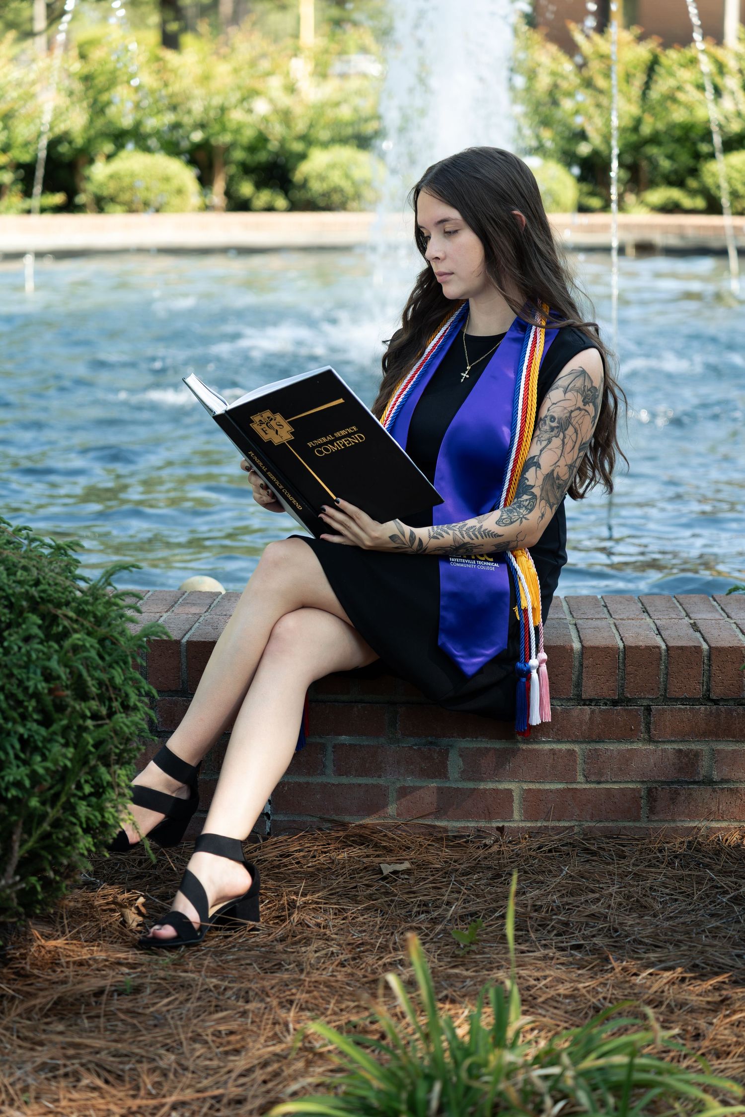 Tattooed female graduate sitting by fountain reading diploma in blue sash and black dress during outdoor graduation photo session.