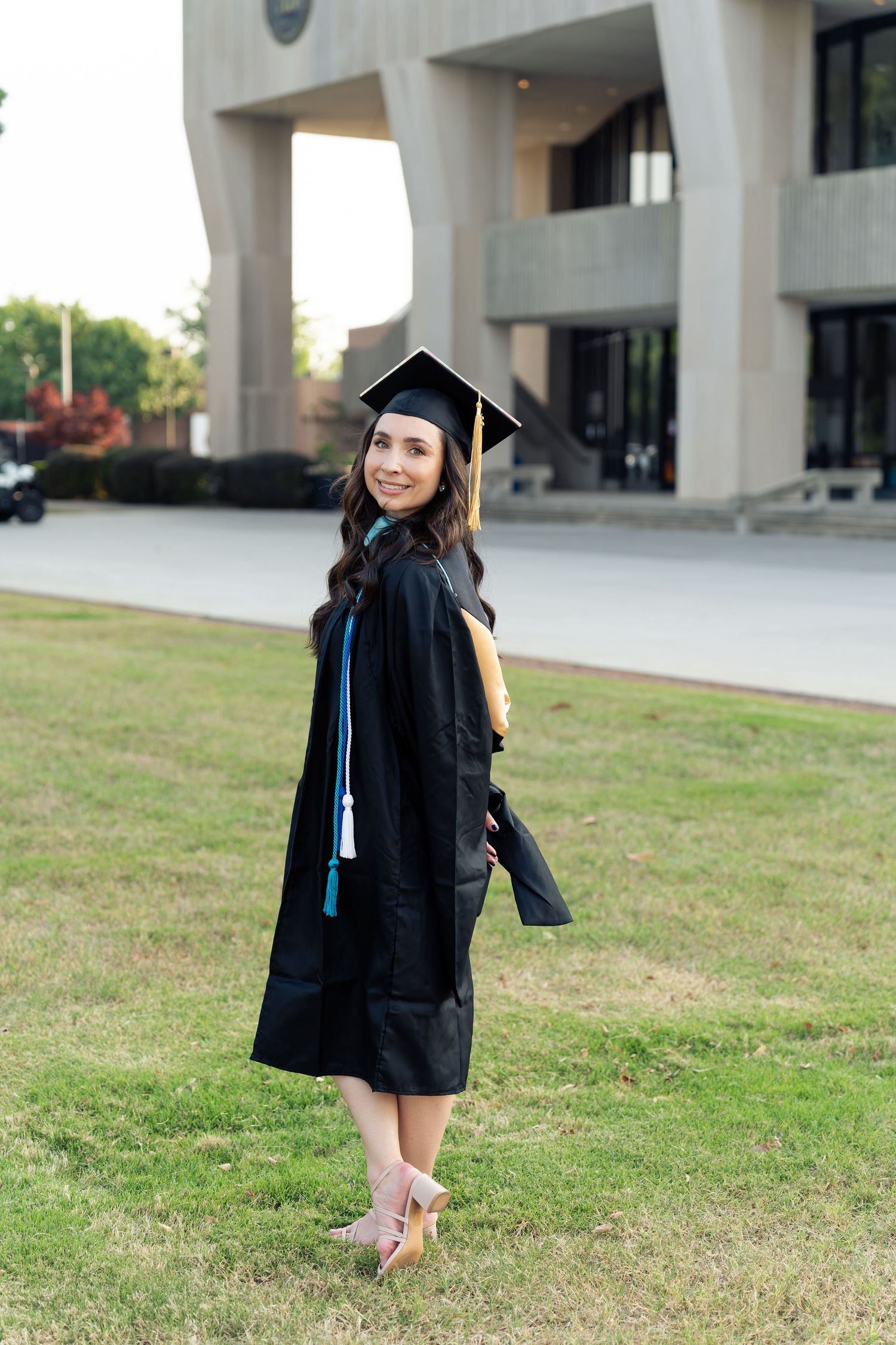 Smiling graduate in black cap and gown holding diploma outdoors on campus lawn.