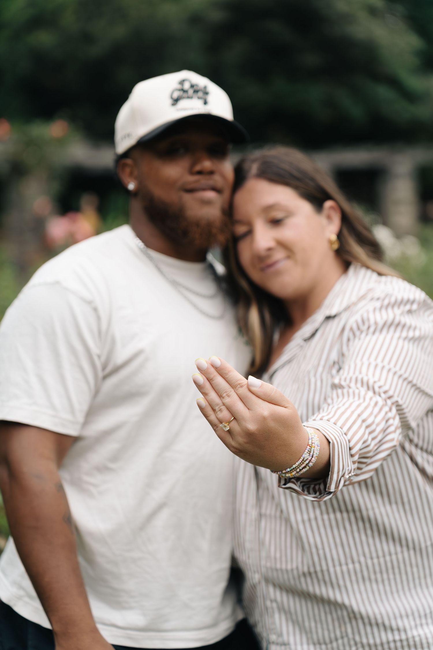 Happy couple showing off engagement ring outdoors, woman extending hand with ring toward camera while leaning on partner.