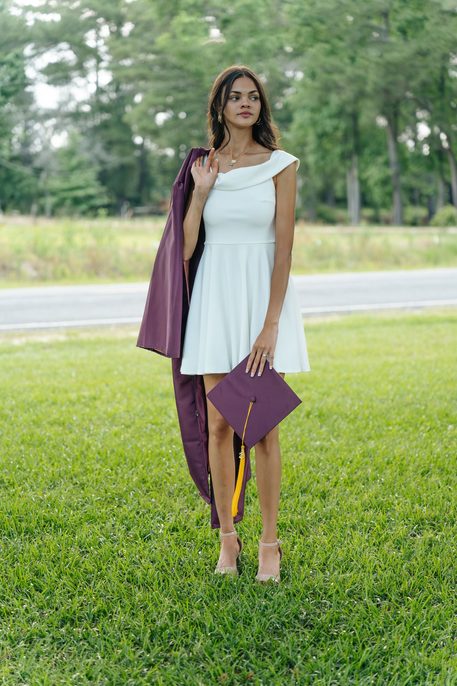 Young woman in white dress holding maroon graduation cap and gown, standing on green grass outdoors.