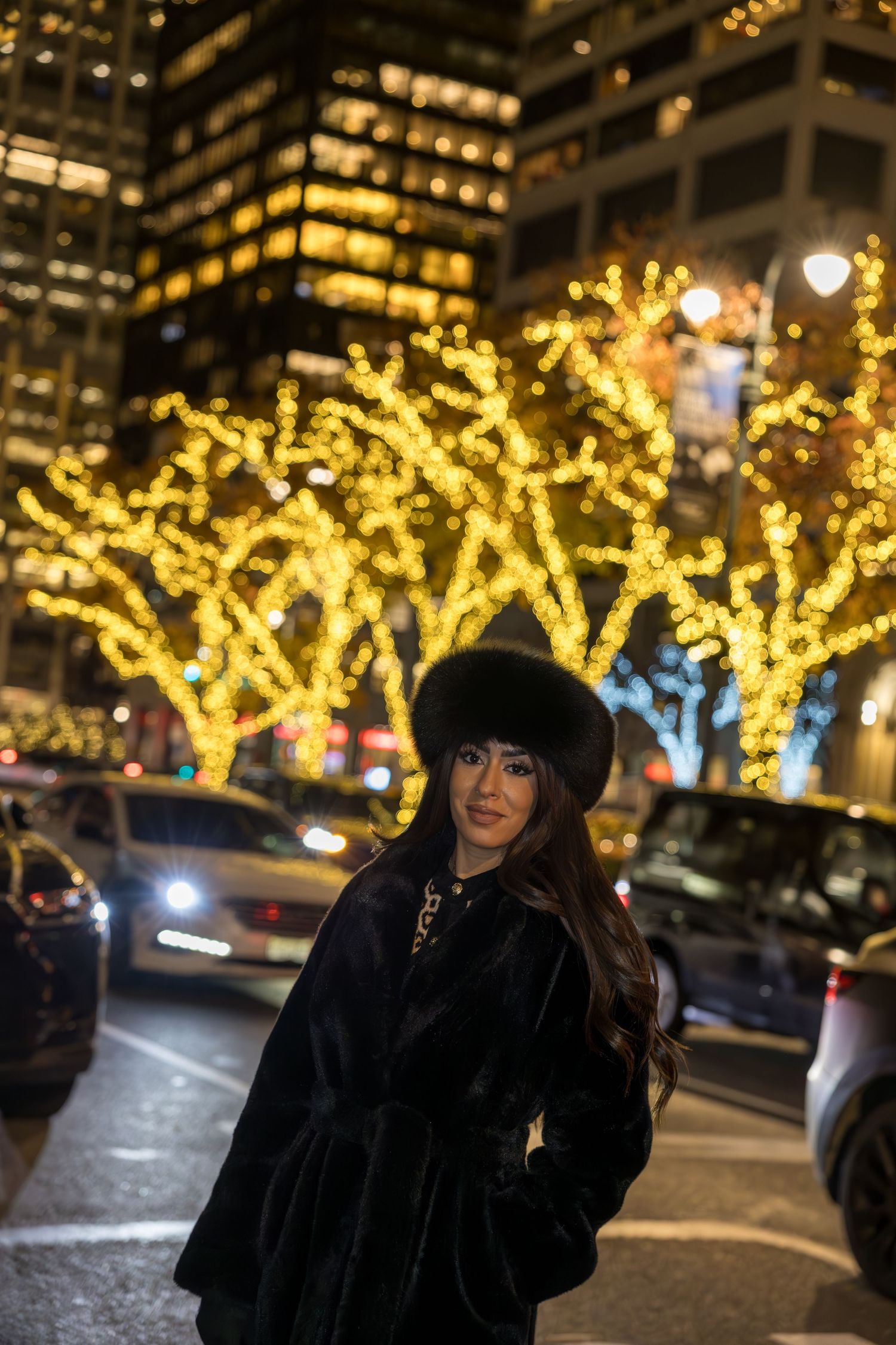 Woman in black fur coat and hat poses on NYC street with golden lit trees at night.