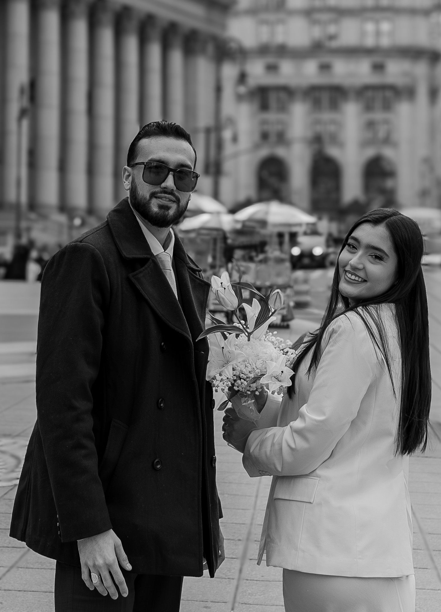 Black and white photo of a smiling couple outside a grand neoclassical building, woman holding white flowers.