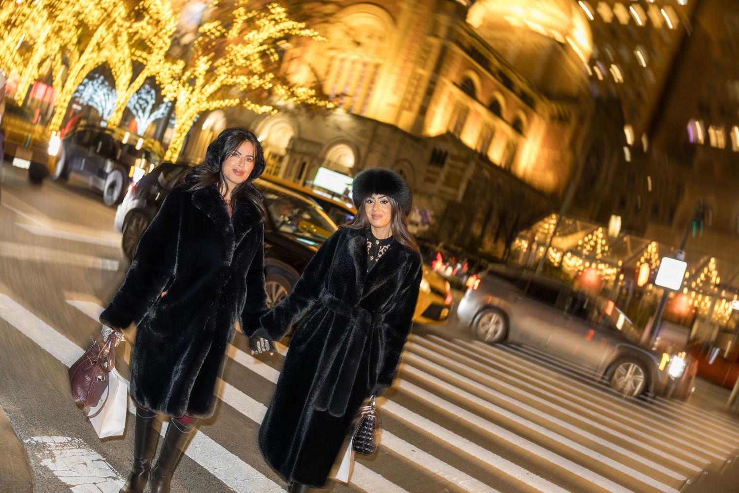 Two women in black fur coats cross a festive night street with golden holiday lights glowing behind them.