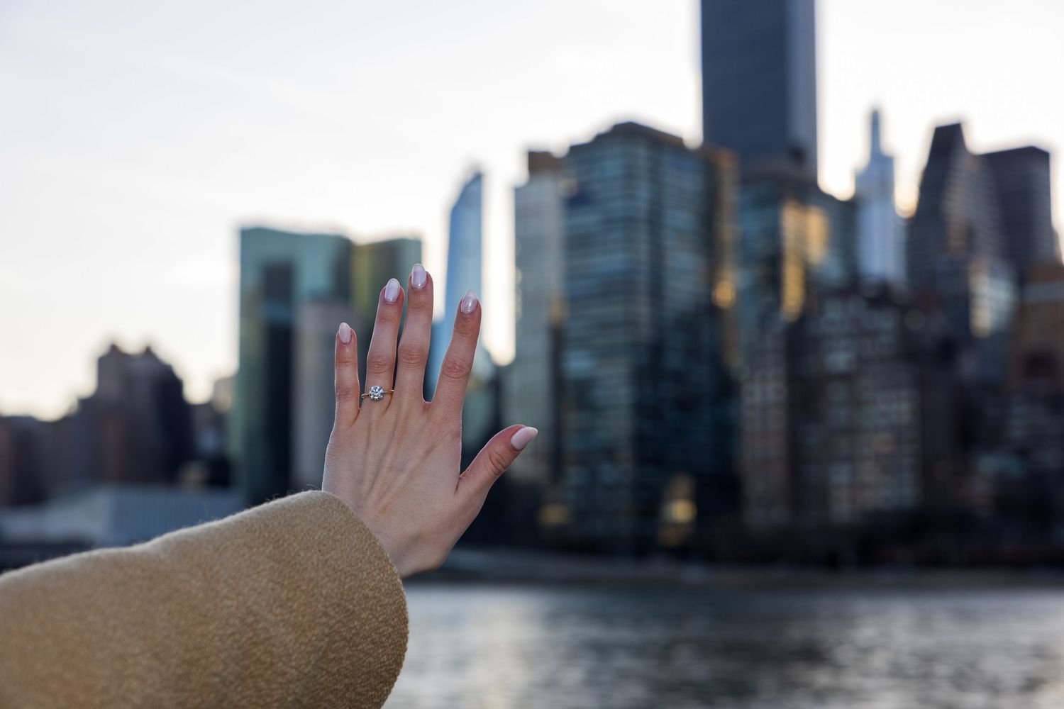 Close-up of a hand displaying an engagement ring with the New York City skyline and river backdrop.
