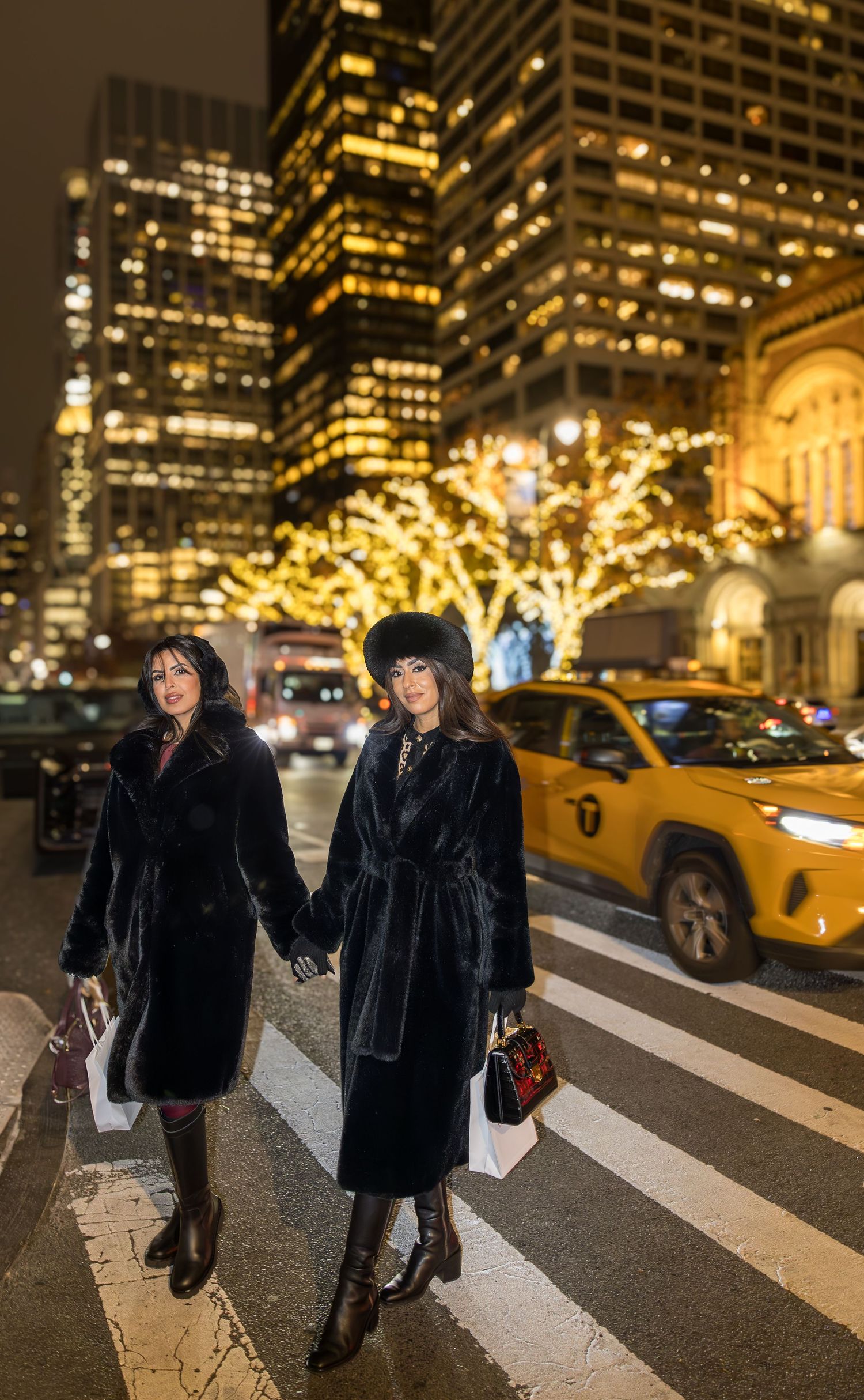 Two stylish women in black fur coats cross a zebra crossing with a yellow taxi and golden lit trees at night.